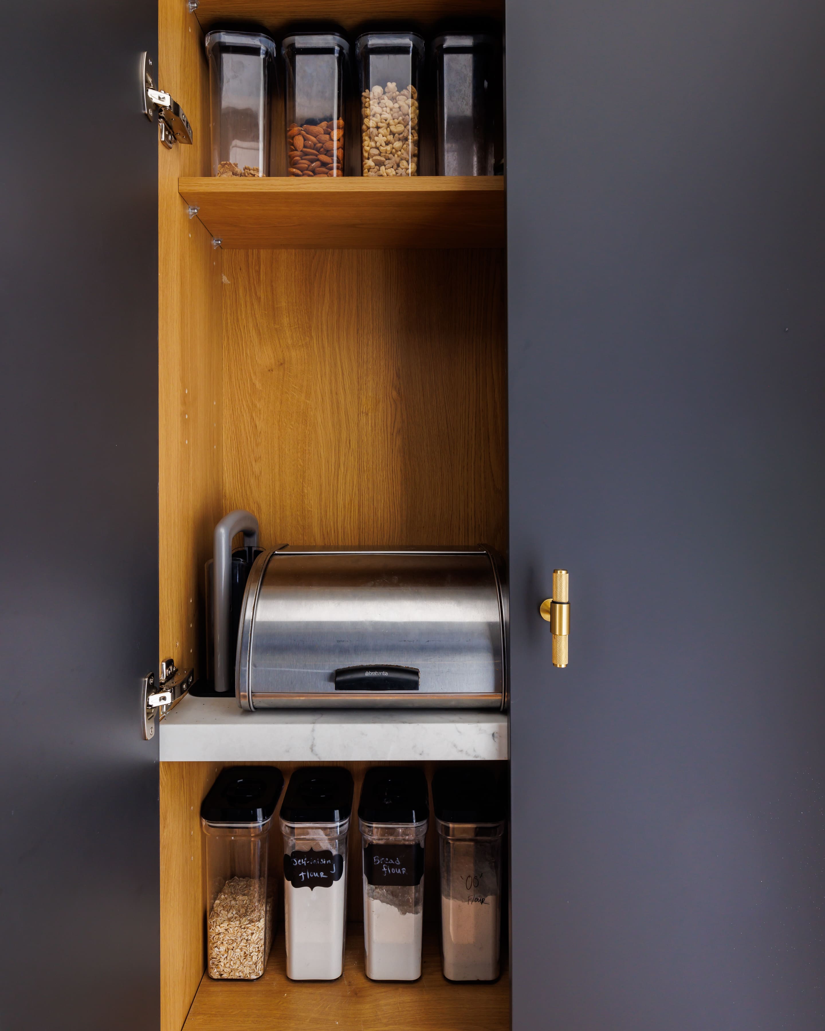 Interior view of a kitchen cupboard featuring a stainless steel bread bin, a marble shelf, and several transparent storage containers, all set against a backdrop of wooden paneling and dark cabinet doors.