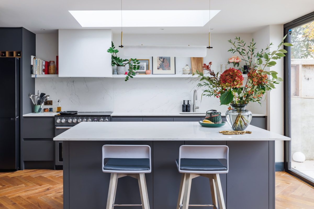 Modern kitchen interior featuring a sleek grey island with two stylish stools, marble countertops, and a skylight above. The space is adorned with vibrant floral arrangements, stylish kitchenware, and tasteful artwork on the walls, creating an inviting and contemporary atmosphere.