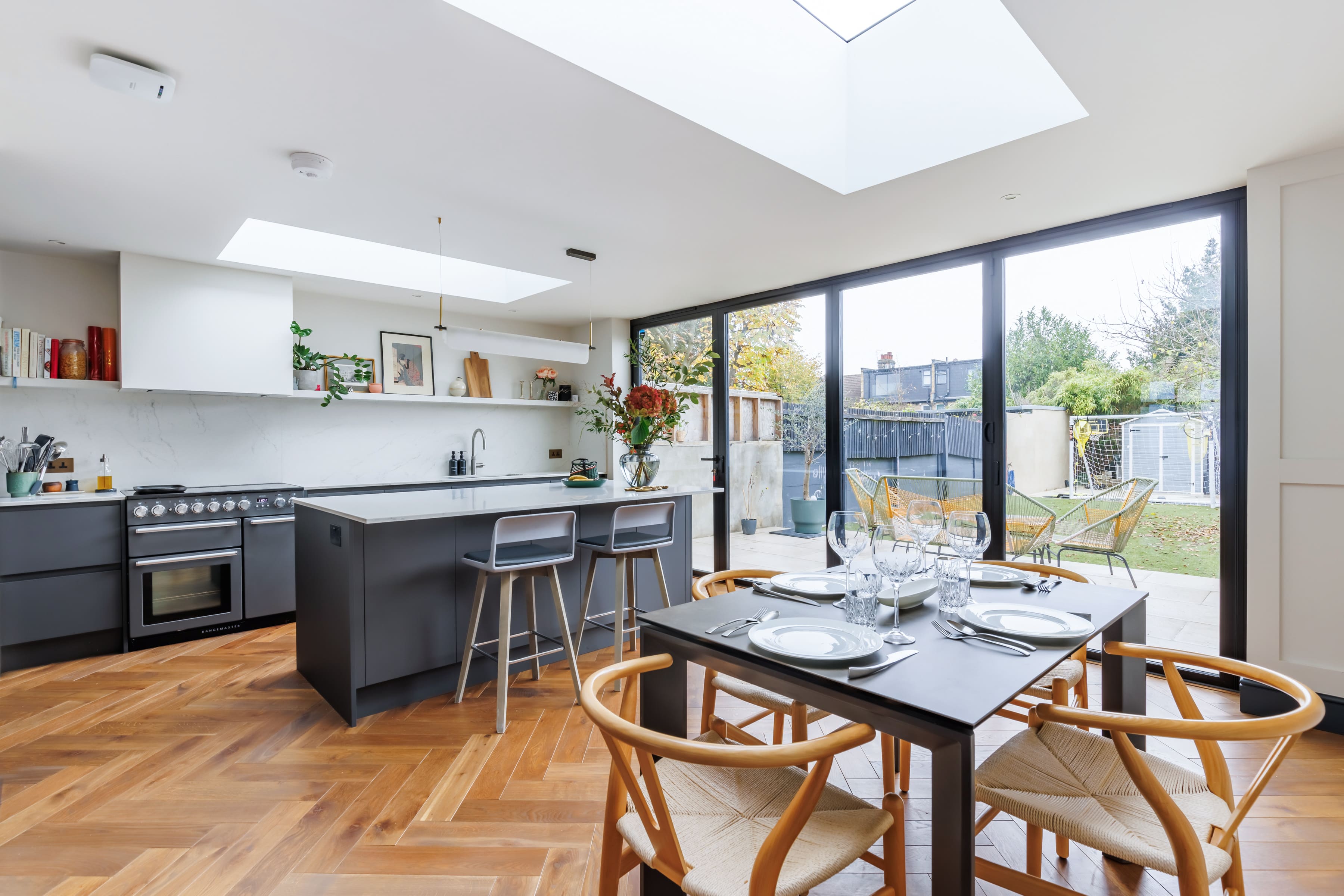 Modern kitchen interior featuring a spacious layout with a central island, stylish bar stools, and a dining table set for a meal. The room is brightened by natural light from a skylight and large glass doors that open to a garden. Elegant decor includes potted plants and a minimalist design with wooden flooring.