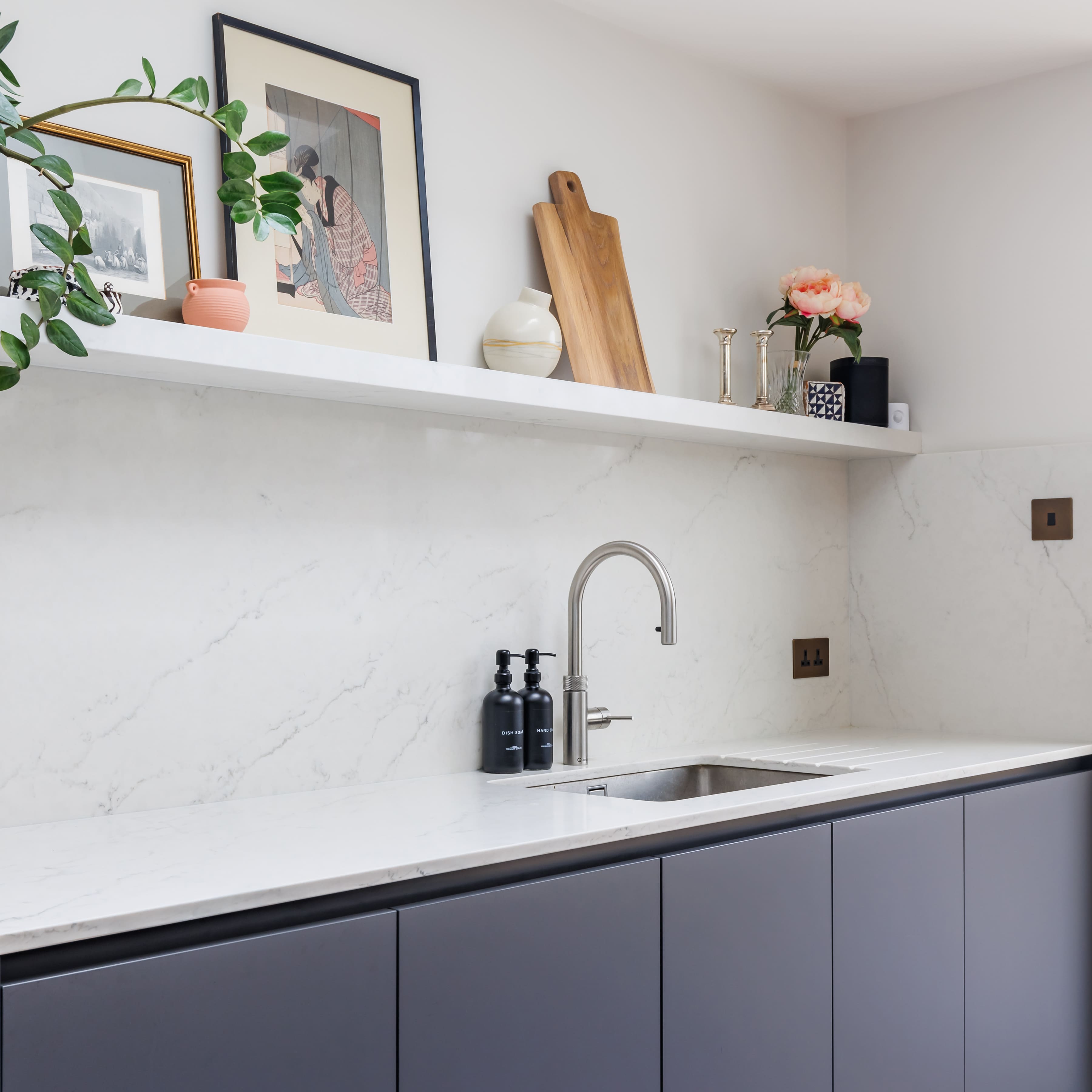 Modern kitchen featuring a sleek marble countertop, stainless steel sink, and stylish black cabinetry. A decorative shelf displays framed art, a wooden cutting board, and floral arrangements, enhancing the contemporary aesthetic.