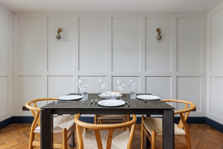 Modern dining room featuring a wooden table set for four, complete with plates, cutlery, and glassware. The space is illuminated by stylish wall sconces against a backdrop of panelled white walls, complemented by herringbone flooring and elegant wooden chairs.