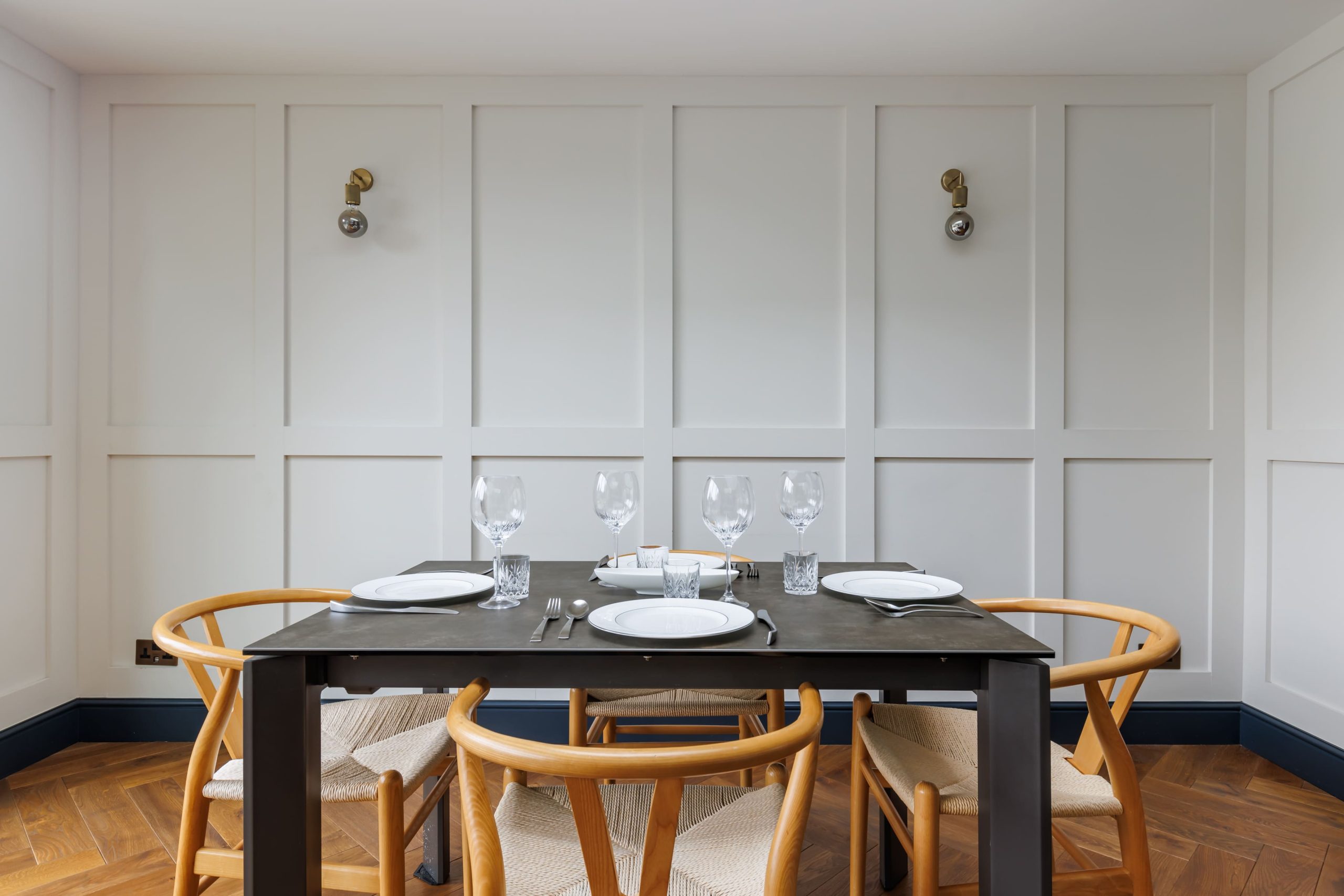Modern dining room featuring a wooden table set for four, complete with plates, cutlery, and glassware. The space is illuminated by stylish wall sconces against a backdrop of panelled white walls, complemented by herringbone flooring and elegant wooden chairs.
