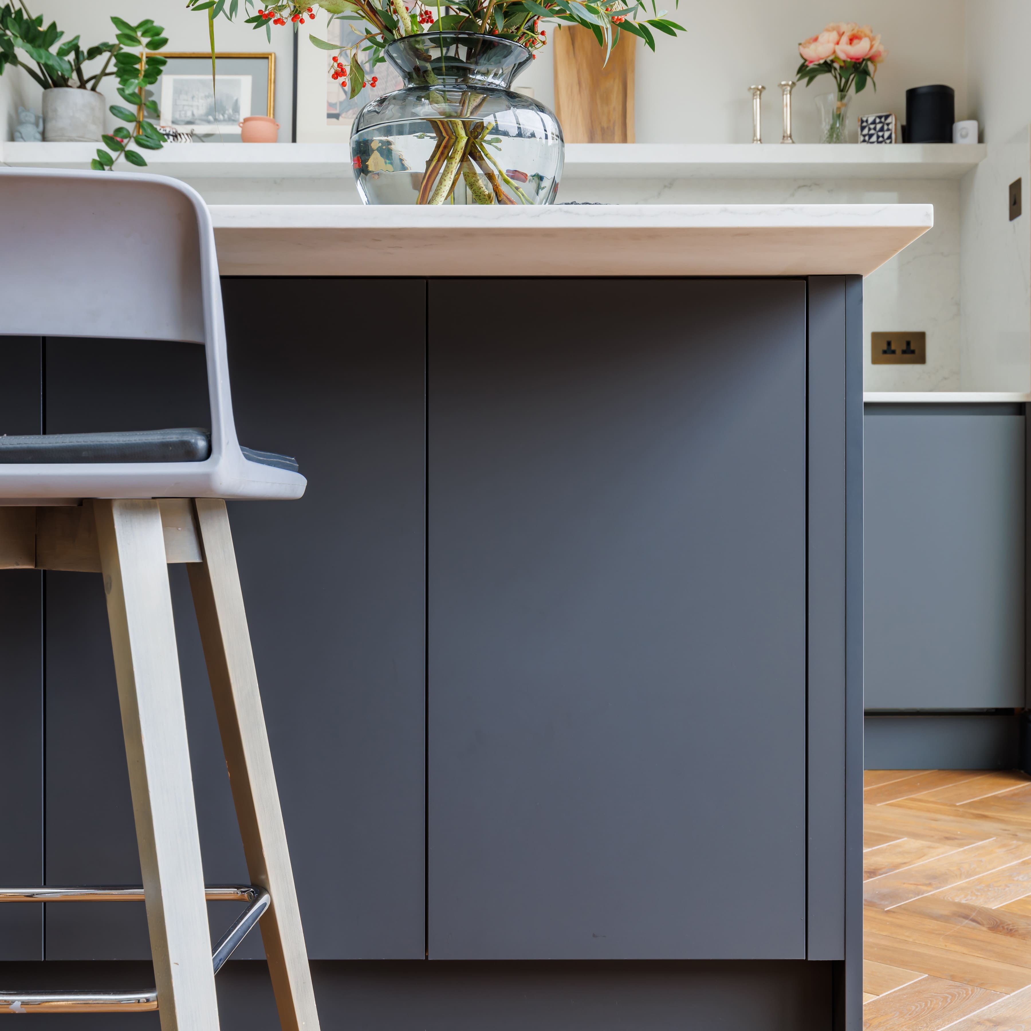 Modern kitchen interior featuring a sleek grey kitchen island with a wooden countertop and a stylish bar stool. A glass vase with decorative plants is placed on the countertop, complementing the contemporary design.
