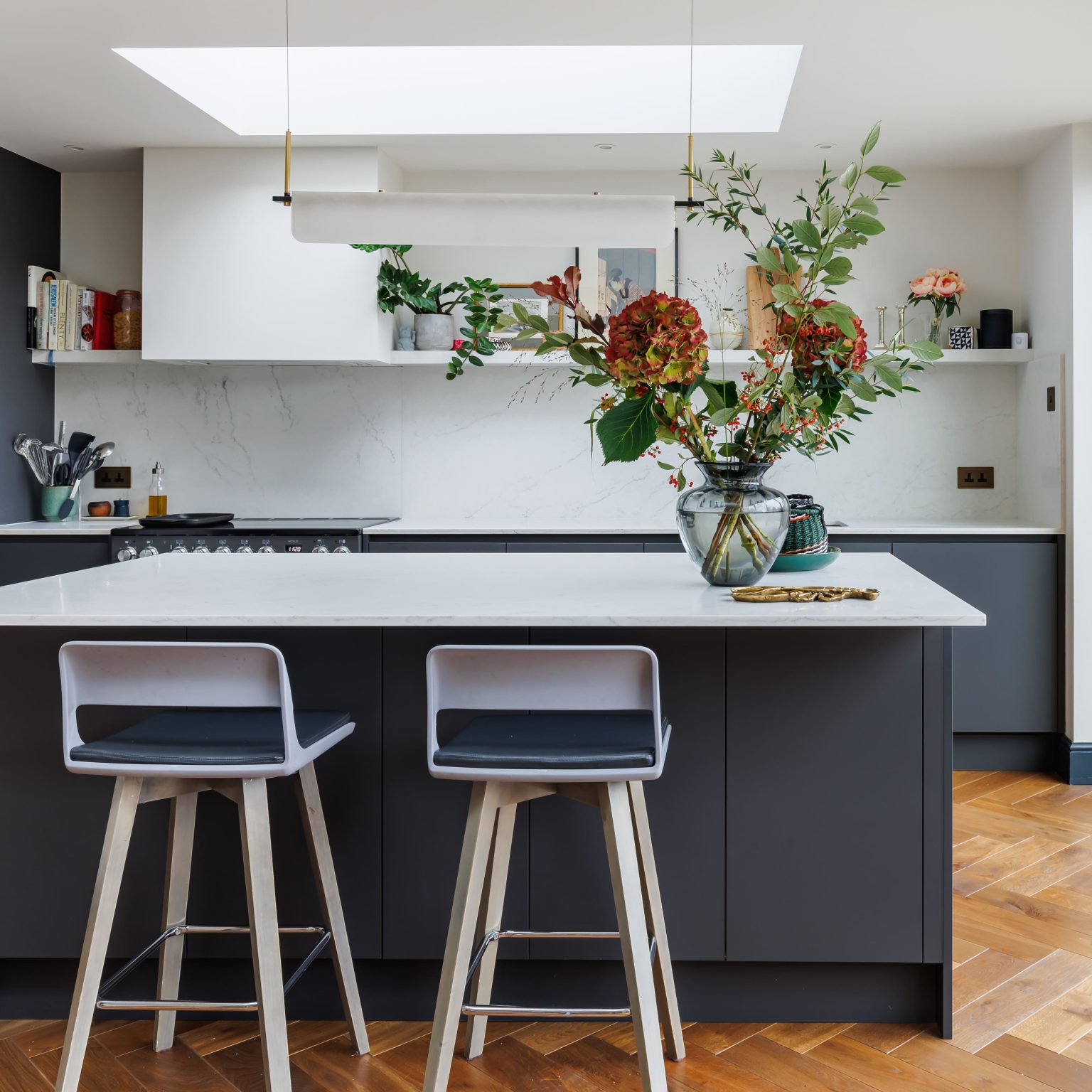 Modern kitchen interior featuring a sleek dark grey island with two bar stools, a white marble worktop, and a decorative vase with vibrant flowers, set against a backdrop of white cabinetry and wooden flooring.