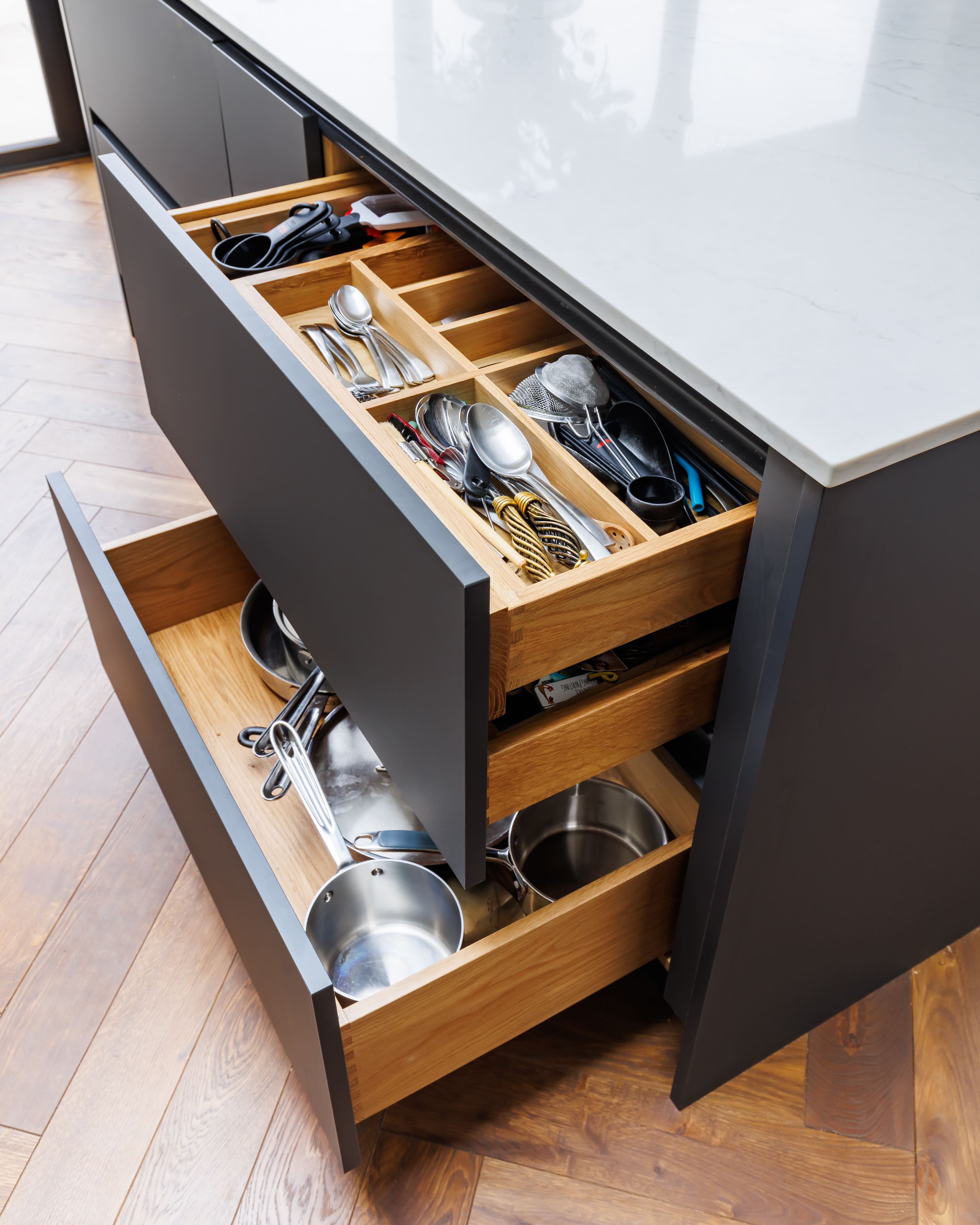 Kitchen drawers showcasing organized cutlery and cookware, including utensils, pots, and pans, in a modern kitchen setting with a marble countertop.