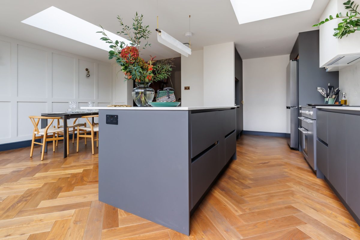 Modern kitchen interior featuring a grey island unit with a floral arrangement on top, hardwood herringbone flooring, and a dining area with a table and chairs. The design includes natural light from a skylight and contemporary kitchen appliances.