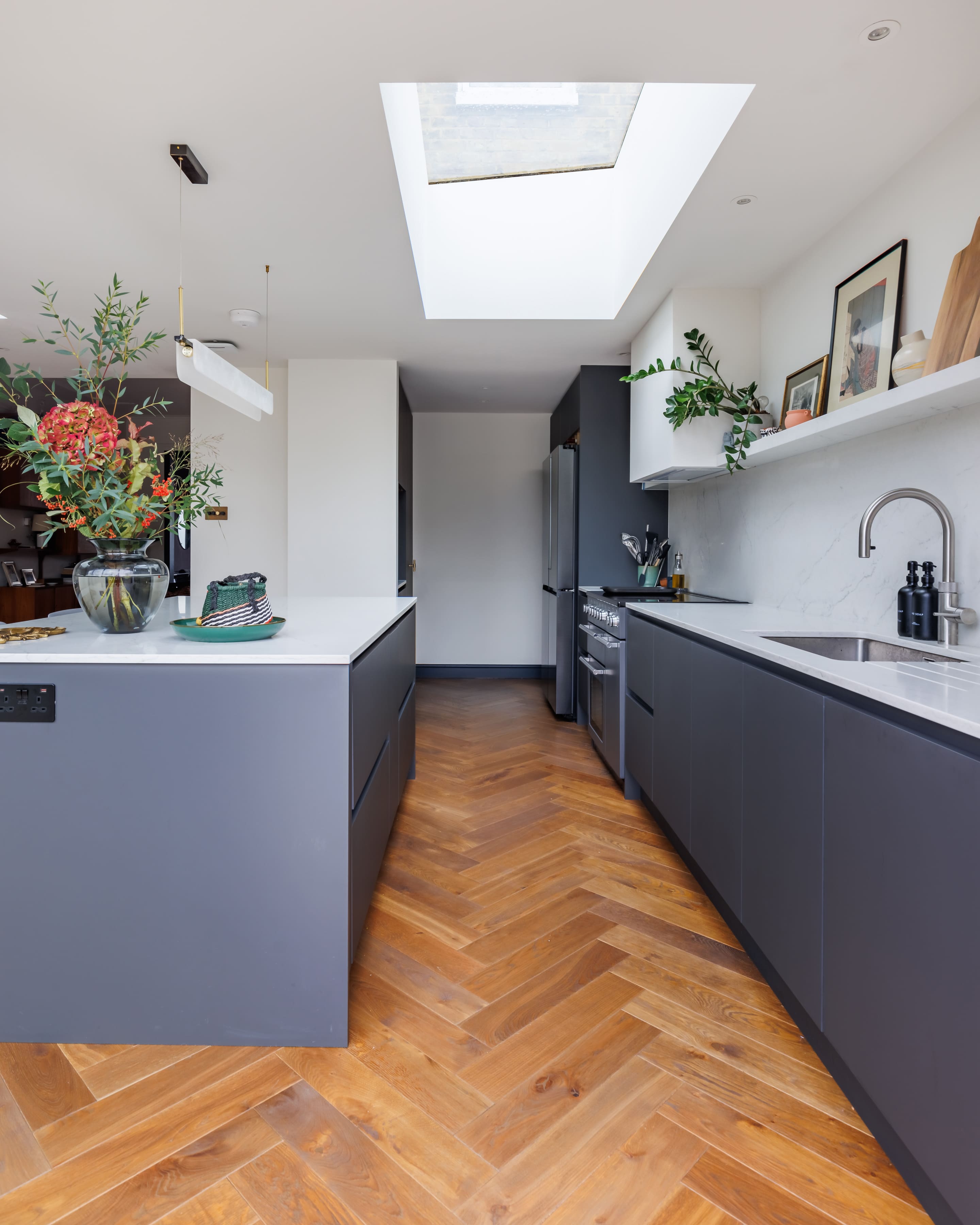 Modern kitchen interior featuring a sleek design with grey cabinetry, a marble countertop, and a herringbone wooden floor. Natural light filters in through a skylight, highlighting decorative plants and kitchen utensils.