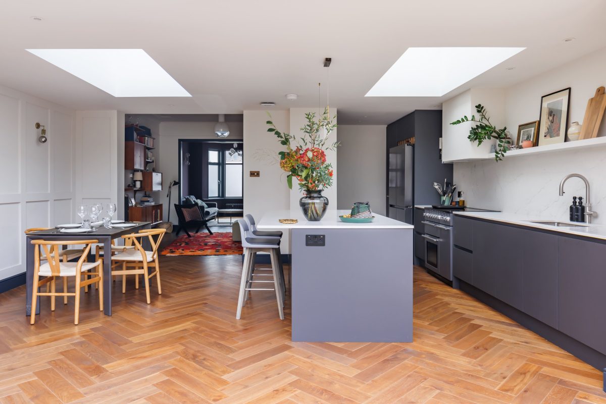Modern open-plan kitchen and dining area featuring a central island, wooden herringbone flooring, and natural light from skylights. The space includes stylish furnishings, fresh flowers in a vase, and a cosy seating area in the background. Ideal for contemporary interior design inspiration.
