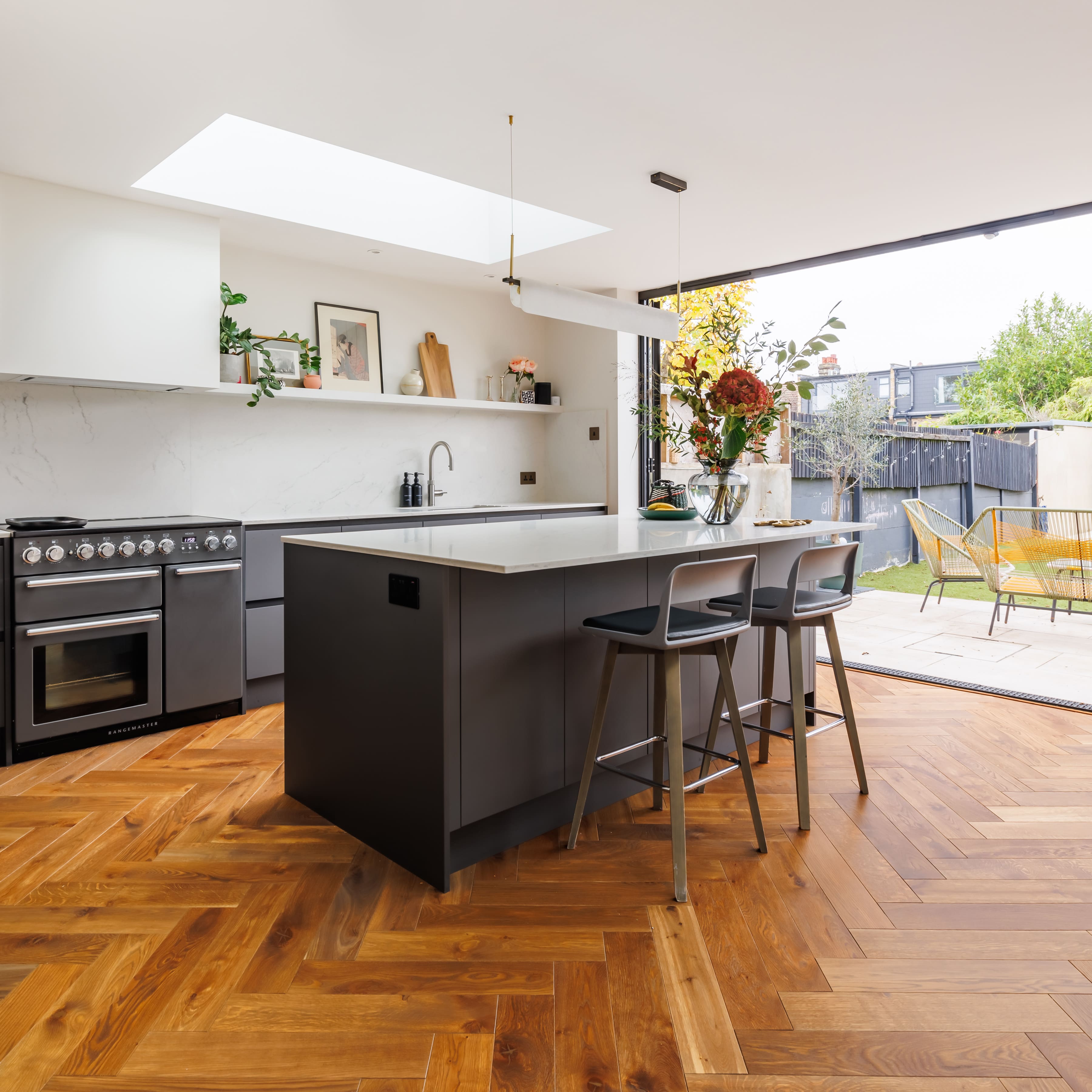 Modern kitchen design featuring a central island with seating, sleek cabinetry, and a stylish oven. Natural light floods the space through large sliding doors, revealing an outdoor patio with garden views. The warm wooden flooring complements the contemporary décor, creating an inviting atmosphere.