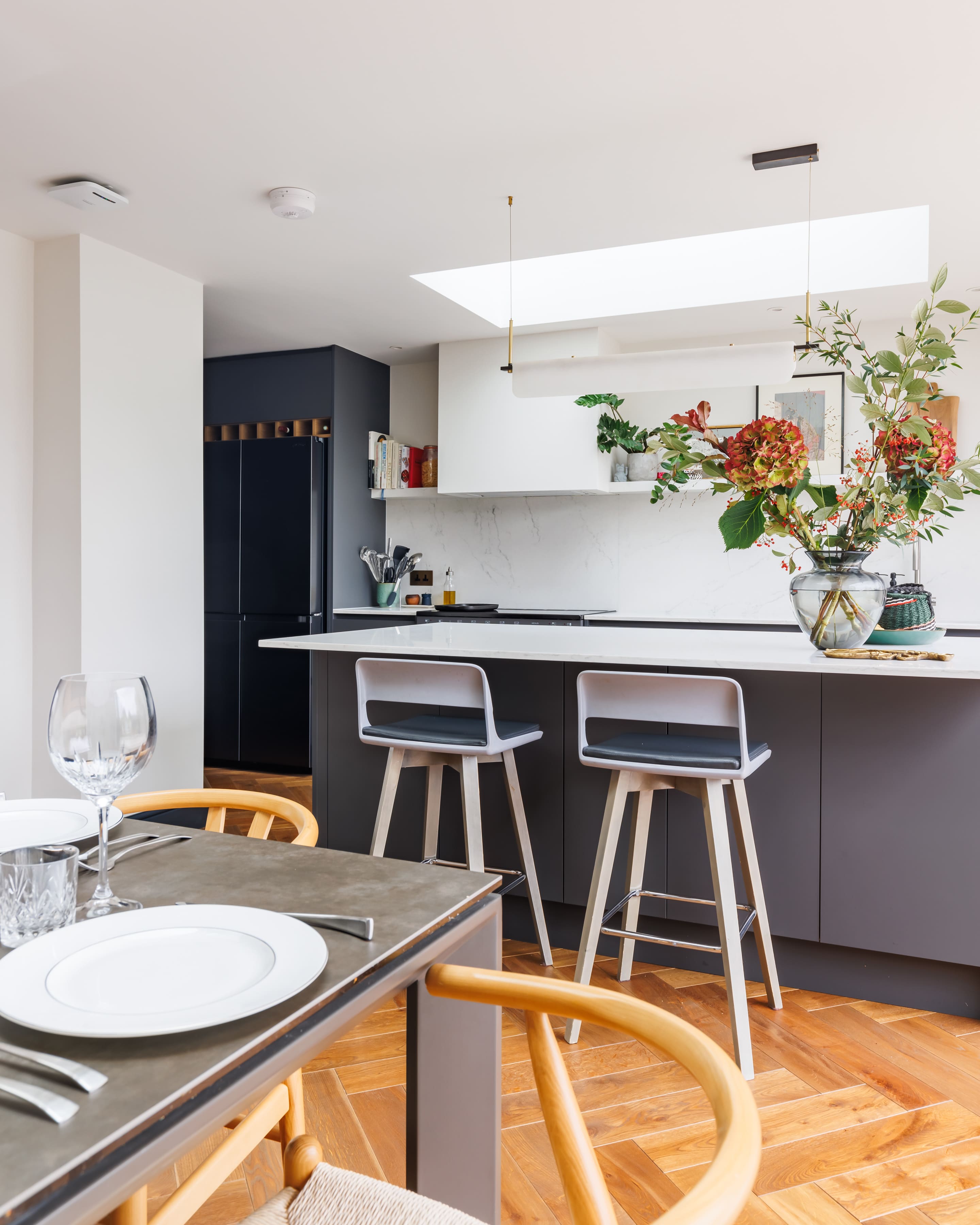 Modern kitchen featuring a sleek design with a marble countertop, minimalist cabinetry, and stylish bar stools. A vibrant floral arrangement sits on the counter, complementing the contemporary decor. Natural light streams in through a skylight, enhancing the open and airy atmosphere.
