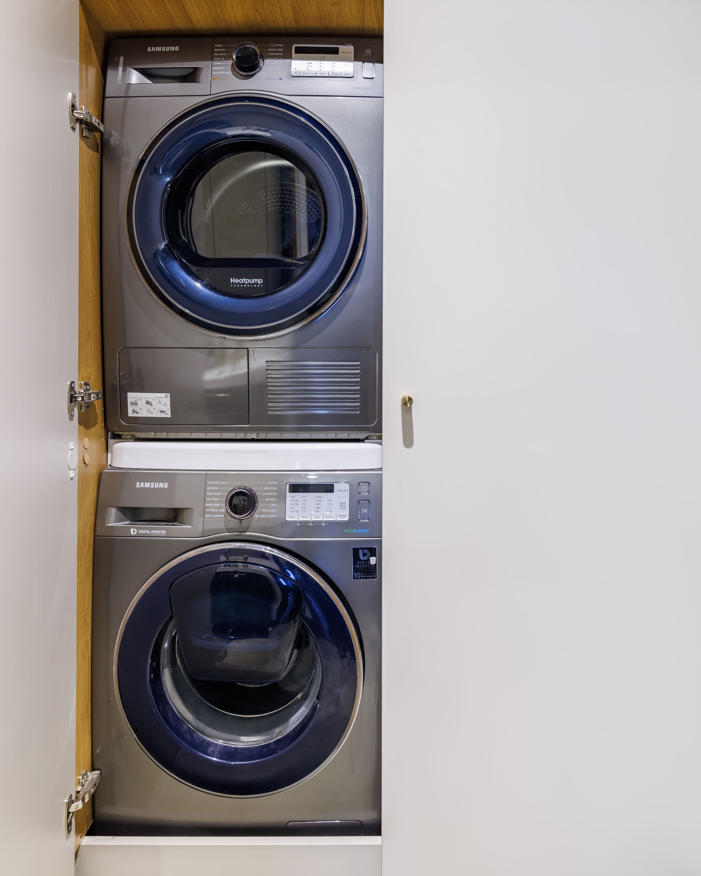 Stacked washer and dryer in a modern laundry space, featuring a Samsung front-loading washing machine and a heat pump tumble dryer, against a minimalist white wall.