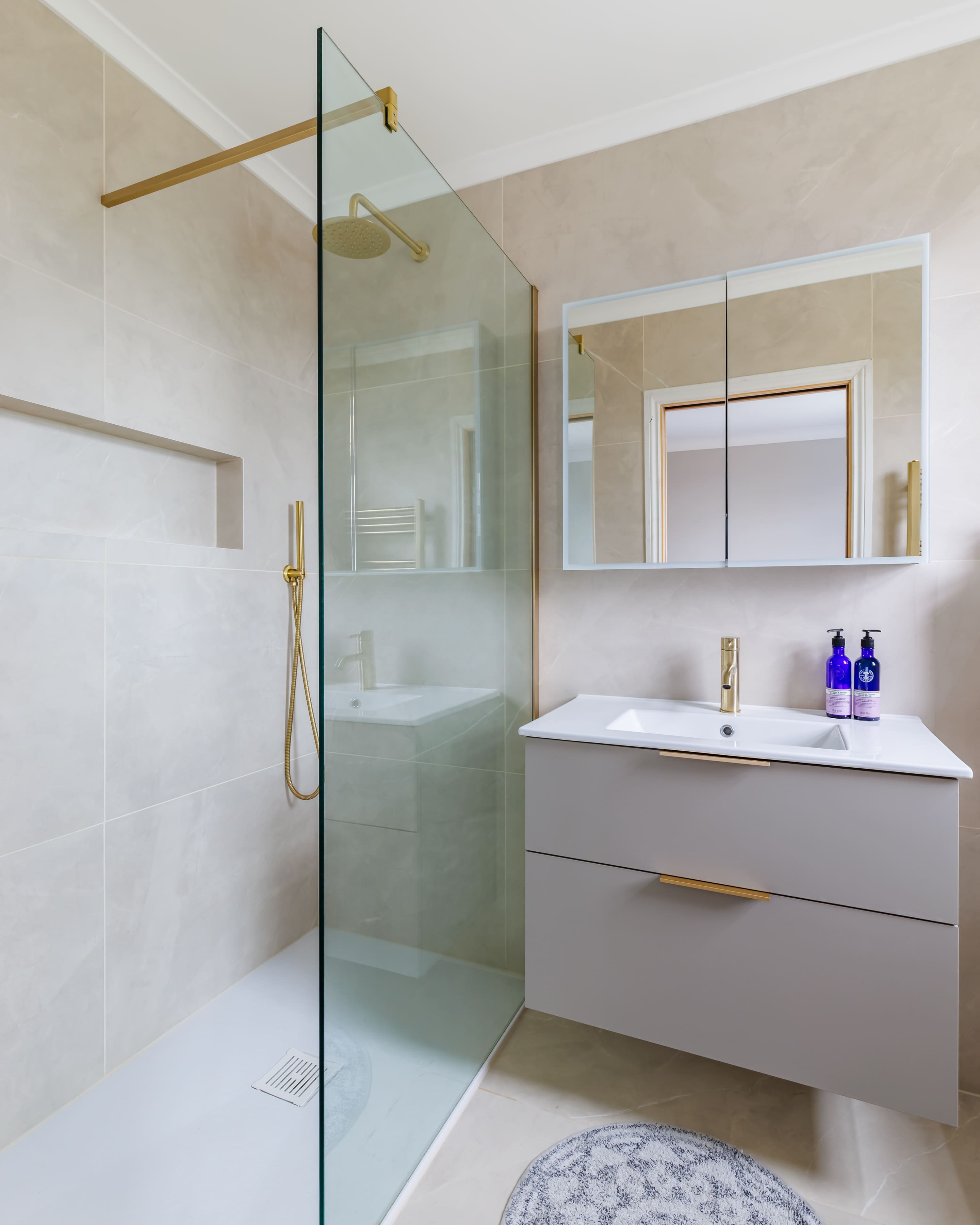 Modern bathroom with a glass shower enclosure, featuring neutral-toned tiled walls, a sleek vanity unit with gold fixtures, and a large mirror. Two blue bottles are placed on the countertop, adding a touch of colour to the minimalist design.