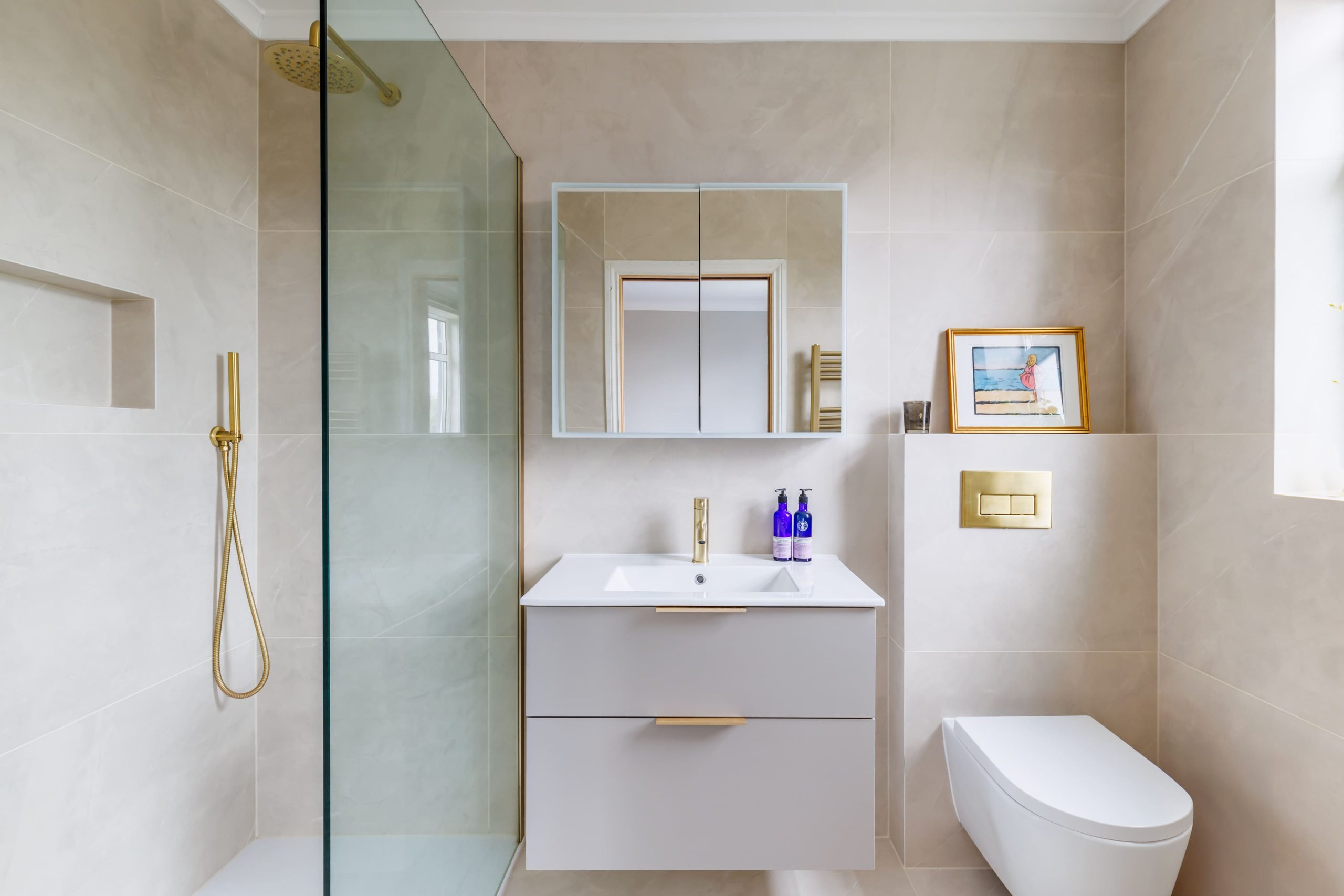Modern bathroom featuring a glass shower enclosure, sleek vanity with a white sink and gold fixtures, large mirror, and minimalist decor. The walls are tiled in neutral tones, complemented by decorative elements.