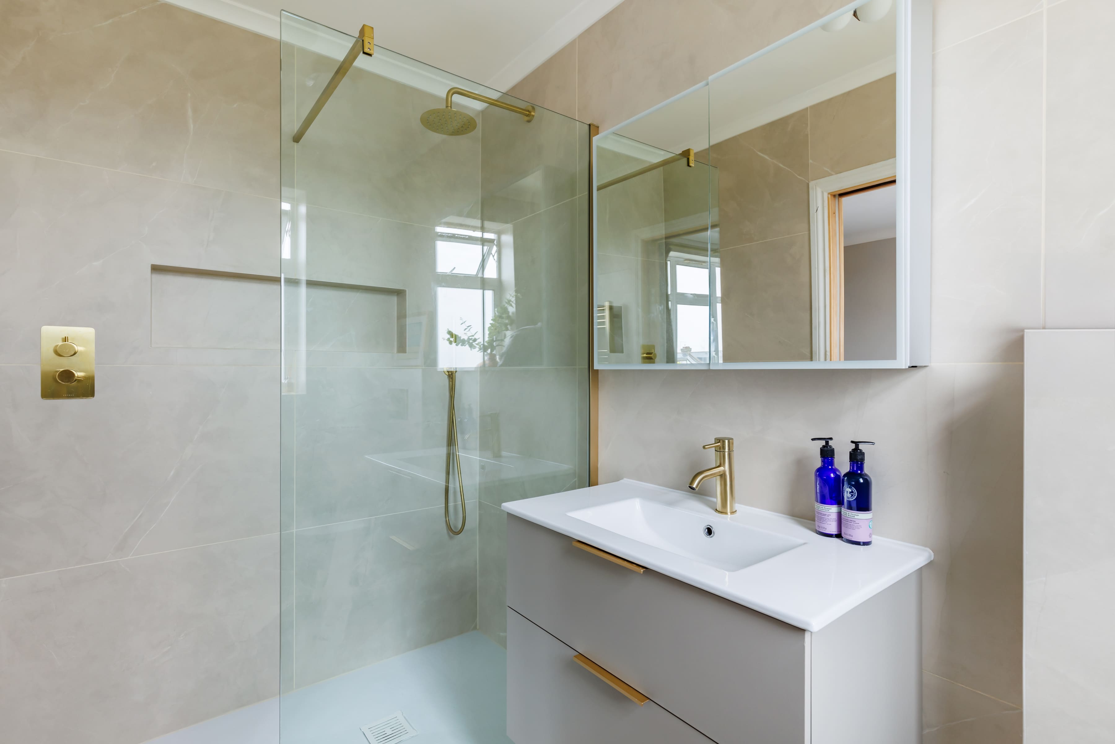 Modern bathroom featuring a glass shower enclosure with a gold showerhead, a sleek grey vanity with a white basin, and decorative blue bottles on the countertop. The walls are tiled in a neutral stone finish, and natural light streams through the windows, enhancing the contemporary design.