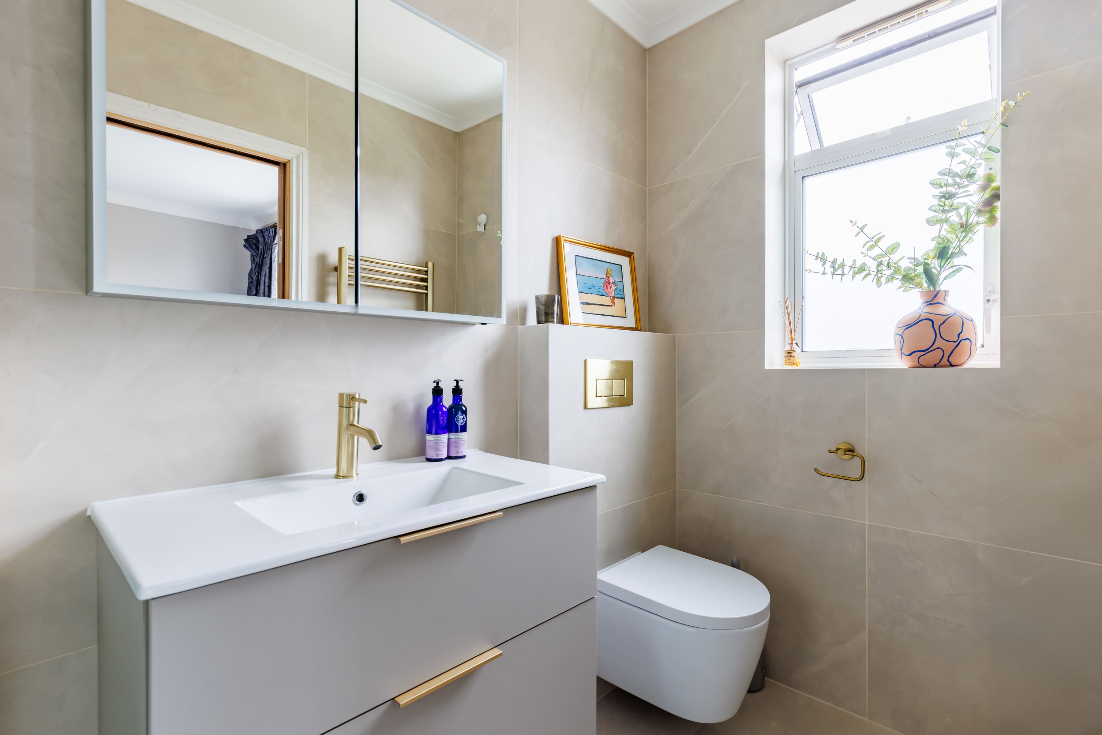 Modern bathroom interior featuring a minimalist design, including a wall-mounted toilet, sleek vanity with a sink, and a large mirror. Natural light streams through a window adorned with a potted plant, enhancing the neutral colour palette and elegant fixtures.