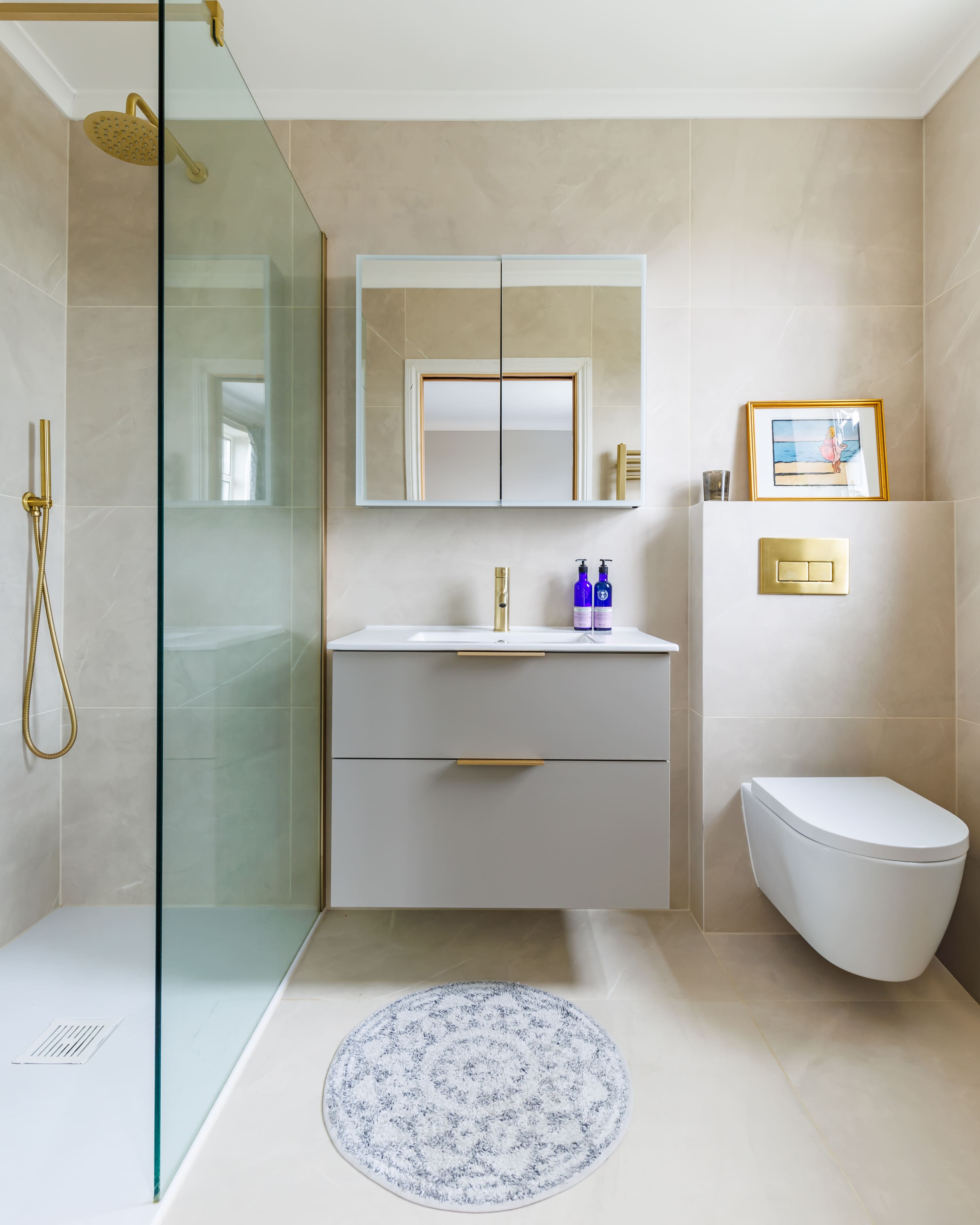 Modern bathroom featuring a walk-in shower with glass partition, a sleek vanity unit with a gold tap and two blue bottles, a wall-mounted toilet, and a stylish mirror. The neutral colour palette and elegant decor create a contemporary and inviting atmosphere.