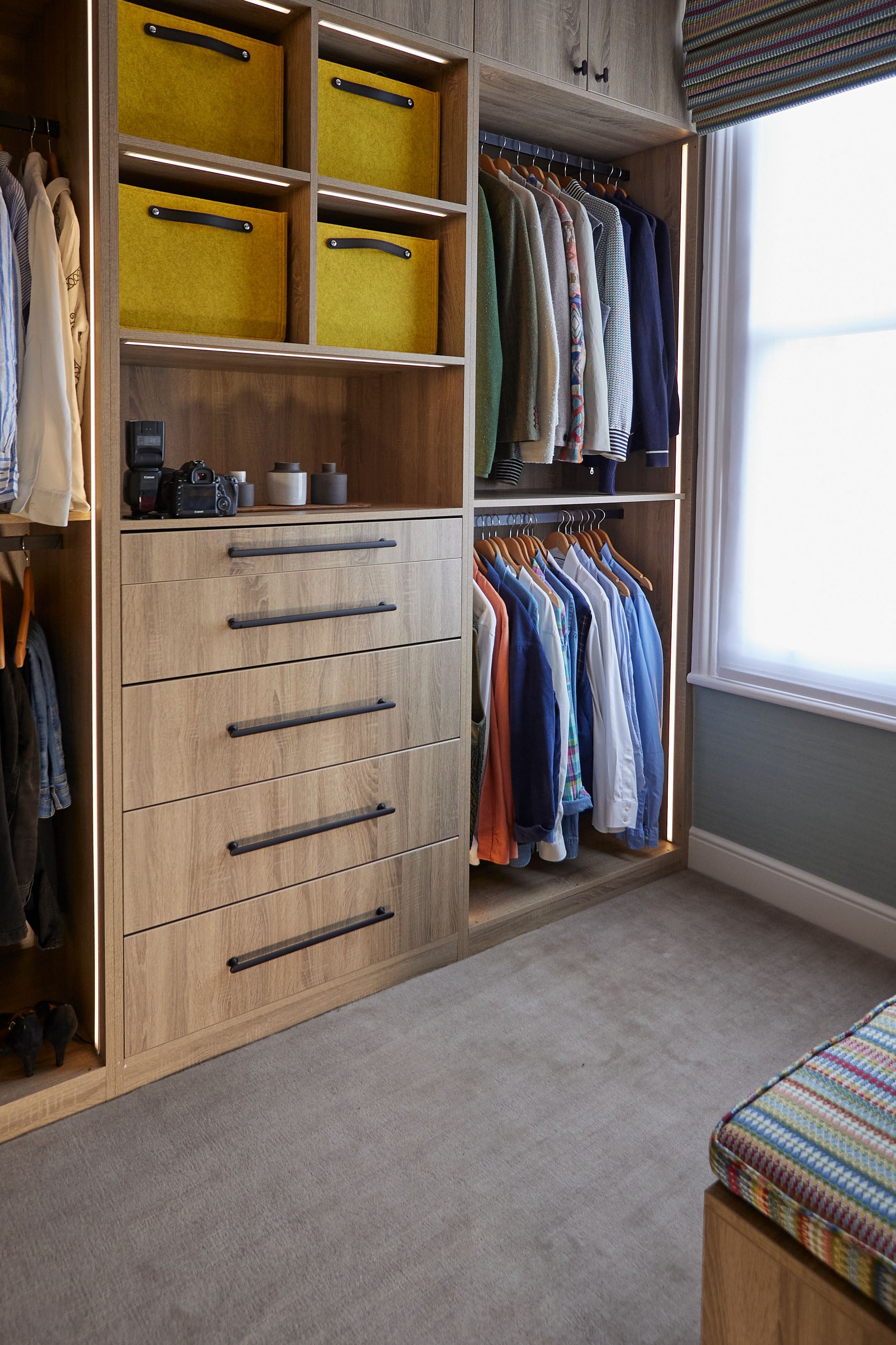 Wooden wardrobe featuring neatly arranged shirts on hangers and a set of drawers. The upper shelf displays personal items, including a camera and decorative containers. The wardrobe is designed for efficient storage and organisation, with a modern aesthetic.