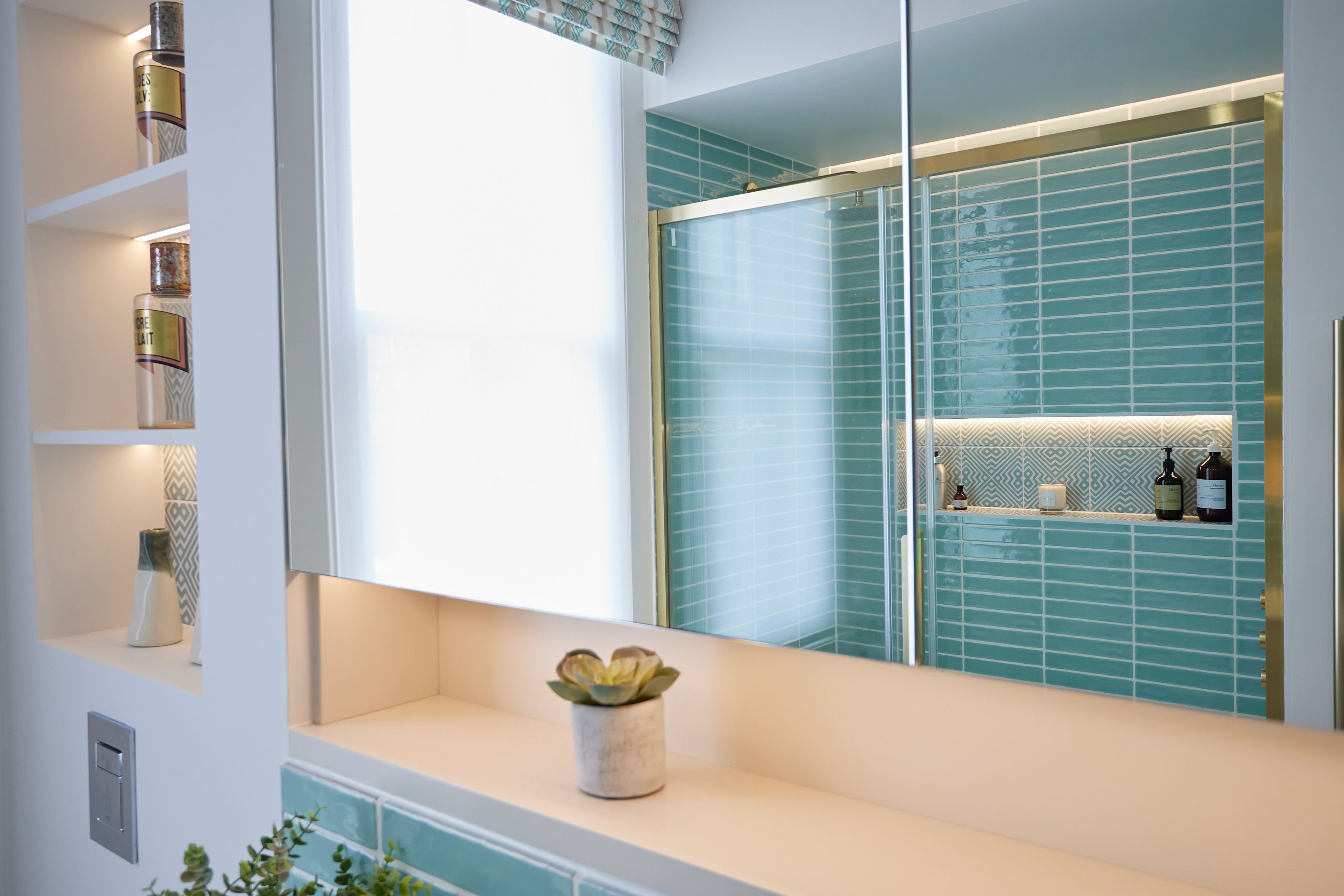 Modern bathroom featuring light blue tiled walls, a large mirror, and a glass shower enclosure. A minimalist shelf displays toiletries and a small potted plant sits on the vanity, enhancing the serene atmosphere. Natural light filters through a window, adding brightness to the space.