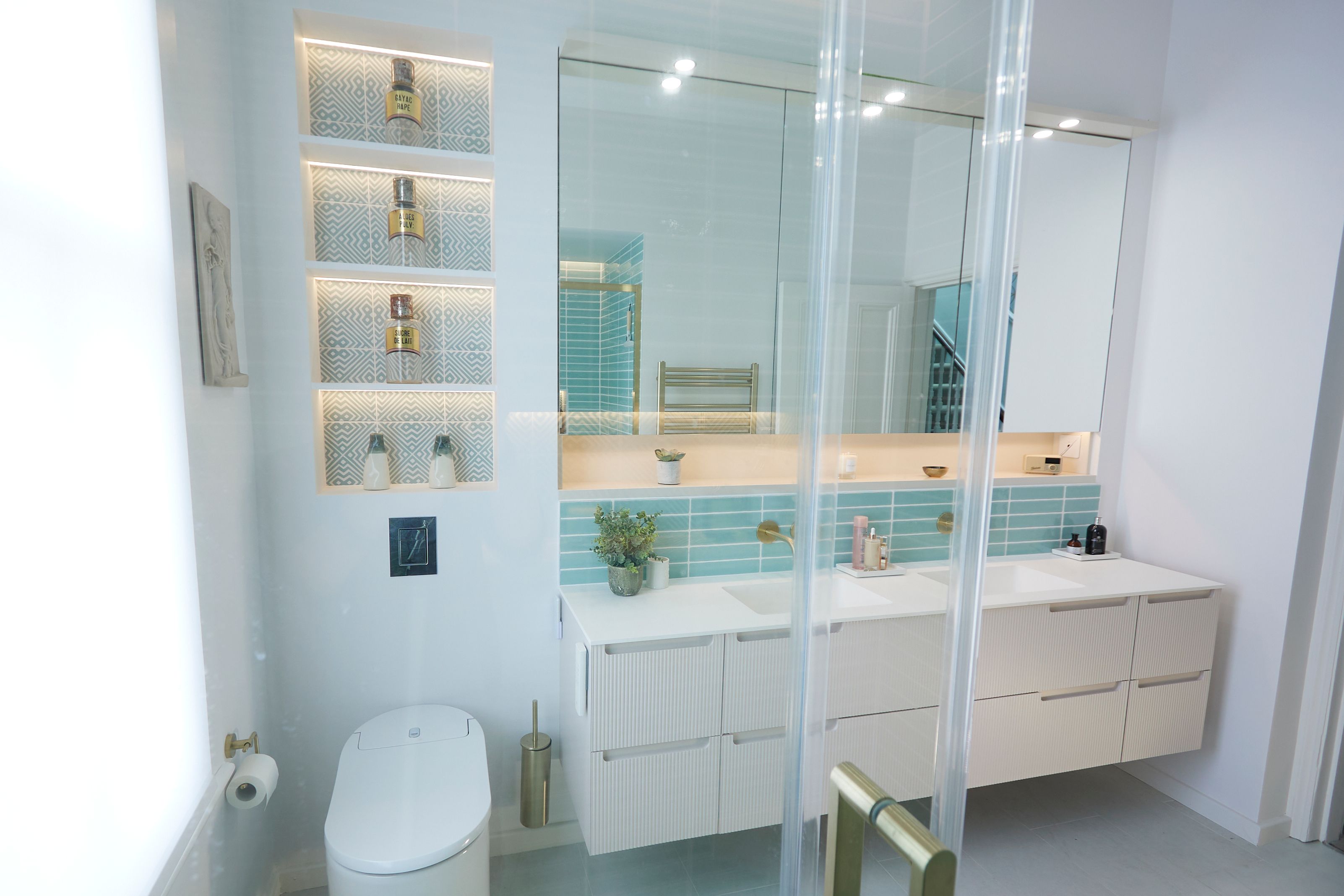 Modern bathroom interior featuring a double vanity with a large mirror, stylish blue tiled backsplash, and elegant lighting. Shelves with decorative items are illuminated, and a sleek toilet is positioned nearby. The design emphasizes a clean, contemporary aesthetic with a focus on functionality and style.