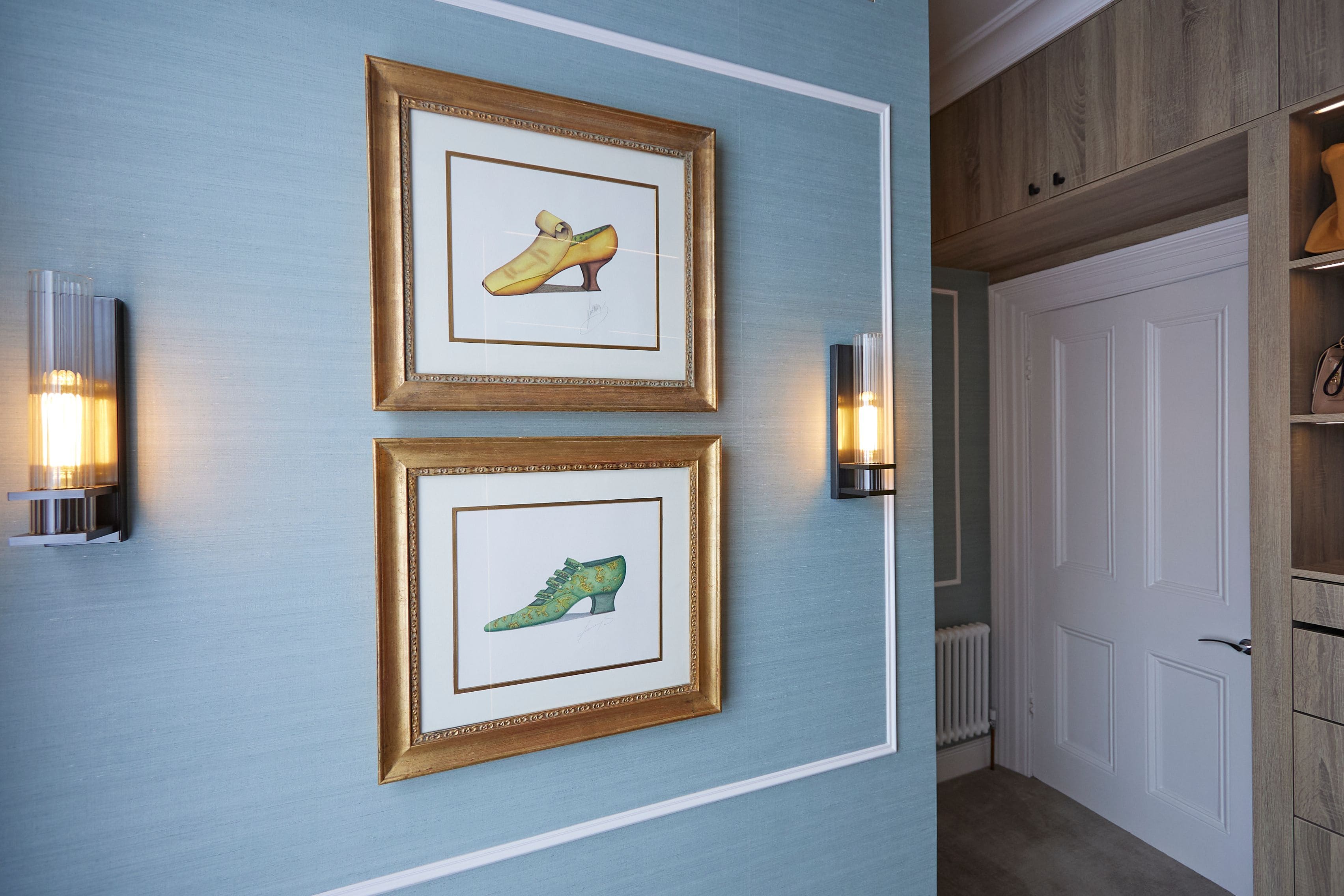 Framed illustrations of stylish footwear, featuring a yellow shoe on top and a green shoe below, displayed on a textured blue wall with contemporary lighting fixtures.