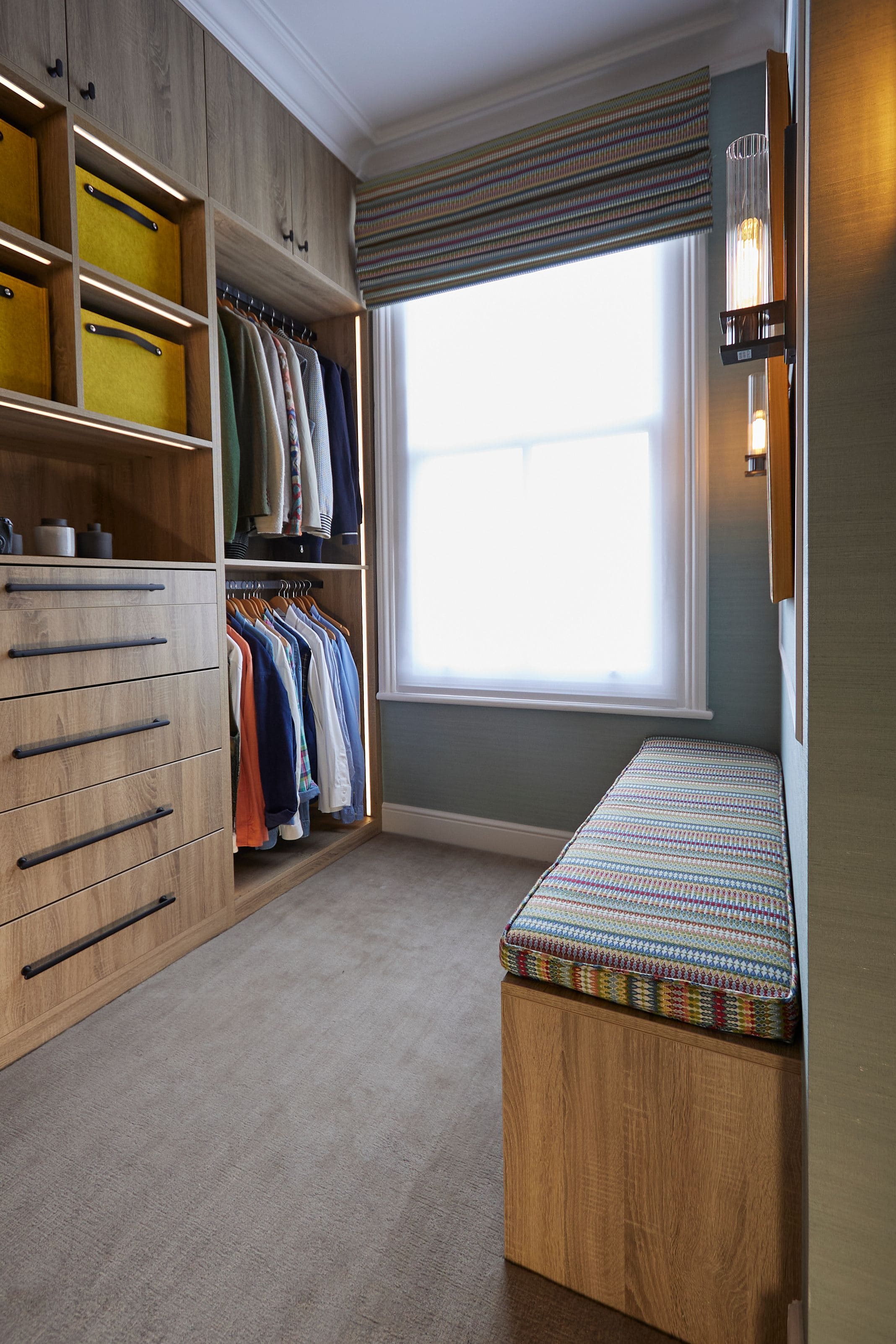 Interior of a stylish wardrobe featuring neatly hung clothes, a wooden drawer unit, and a patterned bench seat by a large window, showcasing an organised and modern storage solution.