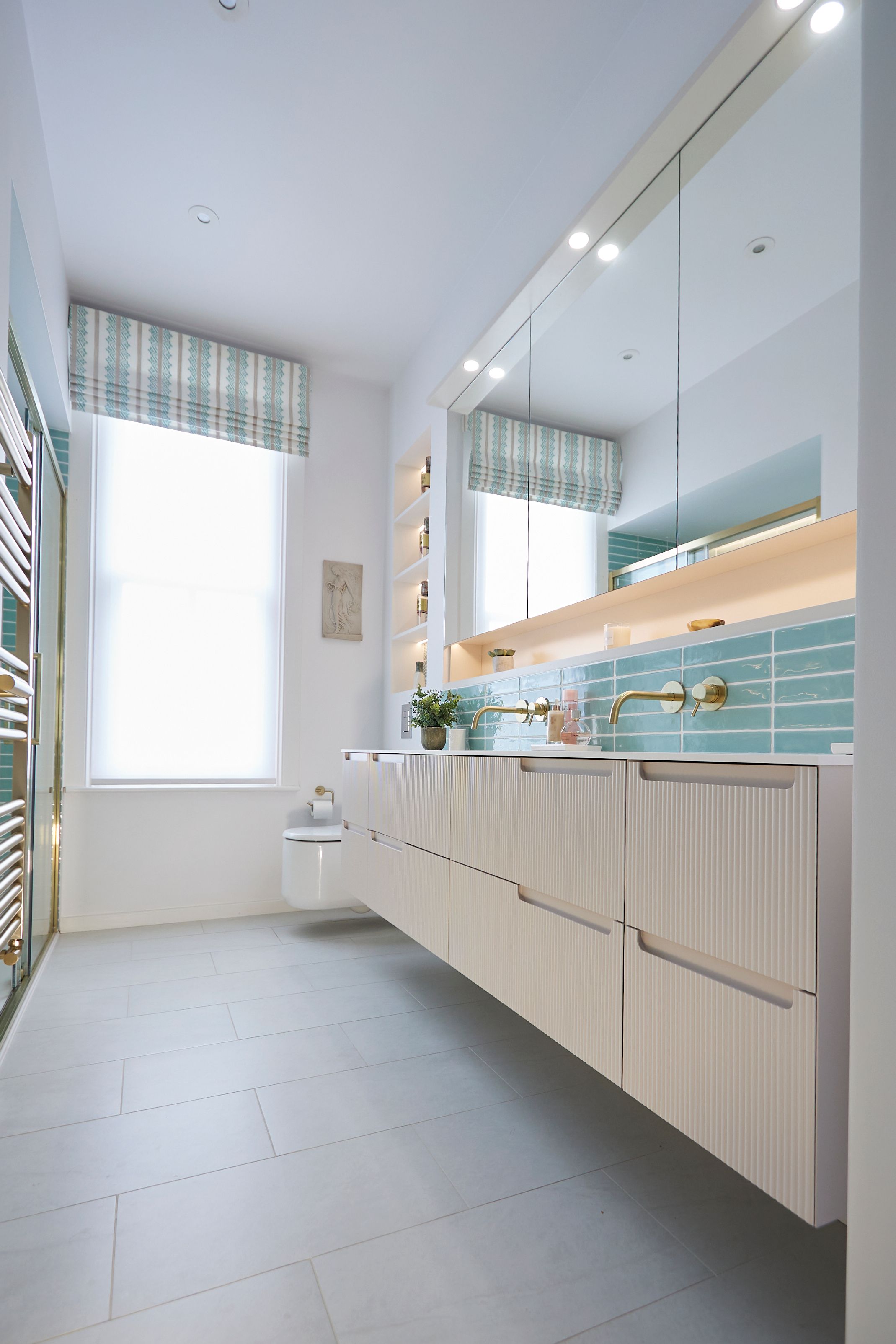 Modern bathroom interior featuring a sleek vanity with textured drawers, a turquoise tiled backsplash, and stylish brass taps. Natural light floods the space through a large window, complemented by a patterned window treatment. Shelving displays decorative items and toiletries, enhancing the contemporary design. A minimalist toilet is positioned discreetly in the corner, contributing to the overall clean aesthetic.