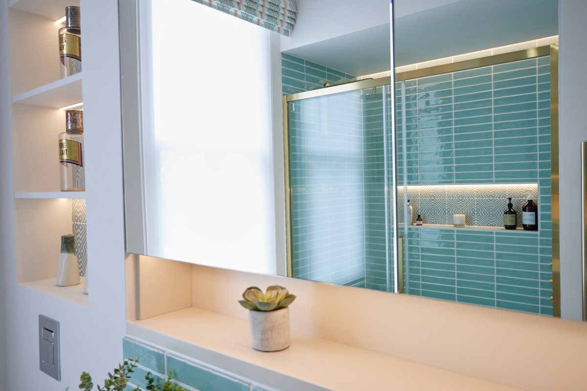 Modern bathroom featuring turquoise tiled walls, a glass shower enclosure, and a sleek vanity with a small potted plant. Natural light filters through a window, enhancing the contemporary design elements and warm tones.