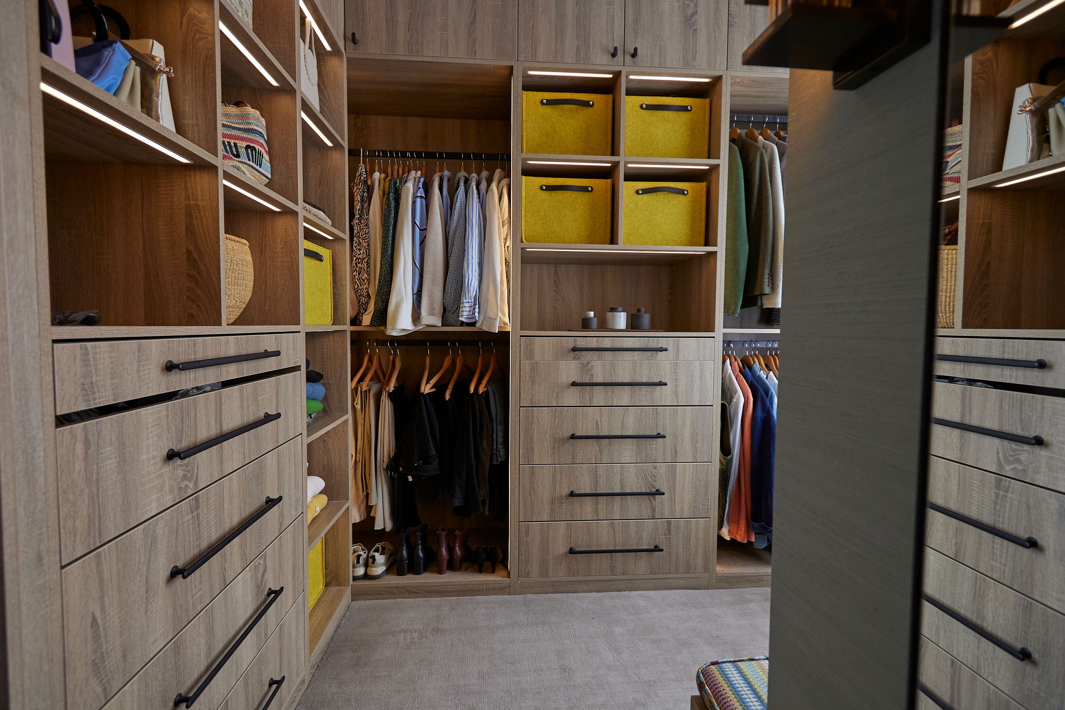 Stylish modern wardrobe with wooden cabinetry, featuring neatly arranged clothing and accessories. The space includes yellow storage boxes, multiple drawers, and hanging sections for garments, creating an organised and aesthetically pleasing storage solution.