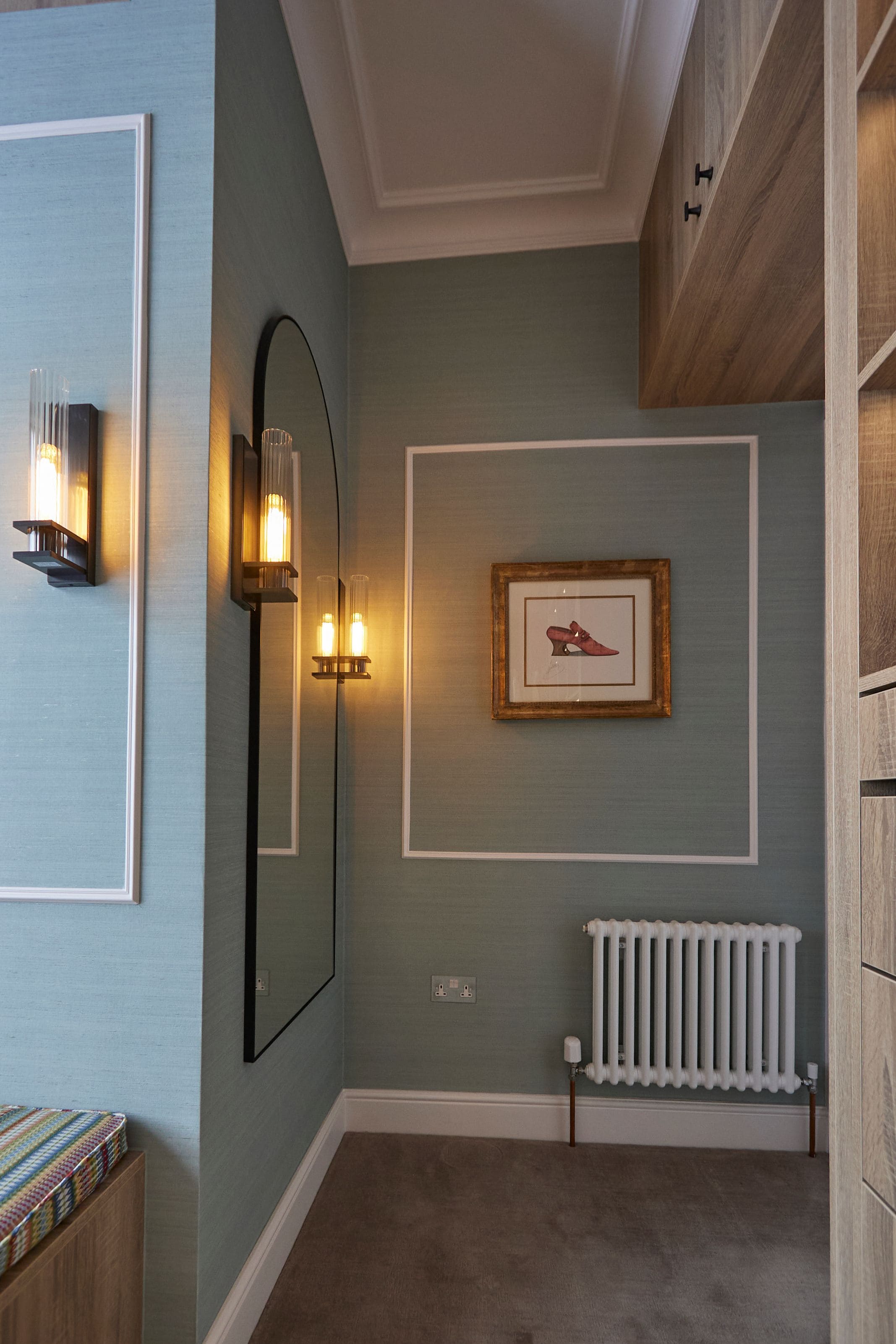 Interior view of a stylish hallway featuring a large mirror, vintage-style wall lights, and a framed artwork of red shoes, complemented by a textured blue wall and a modern radiator.