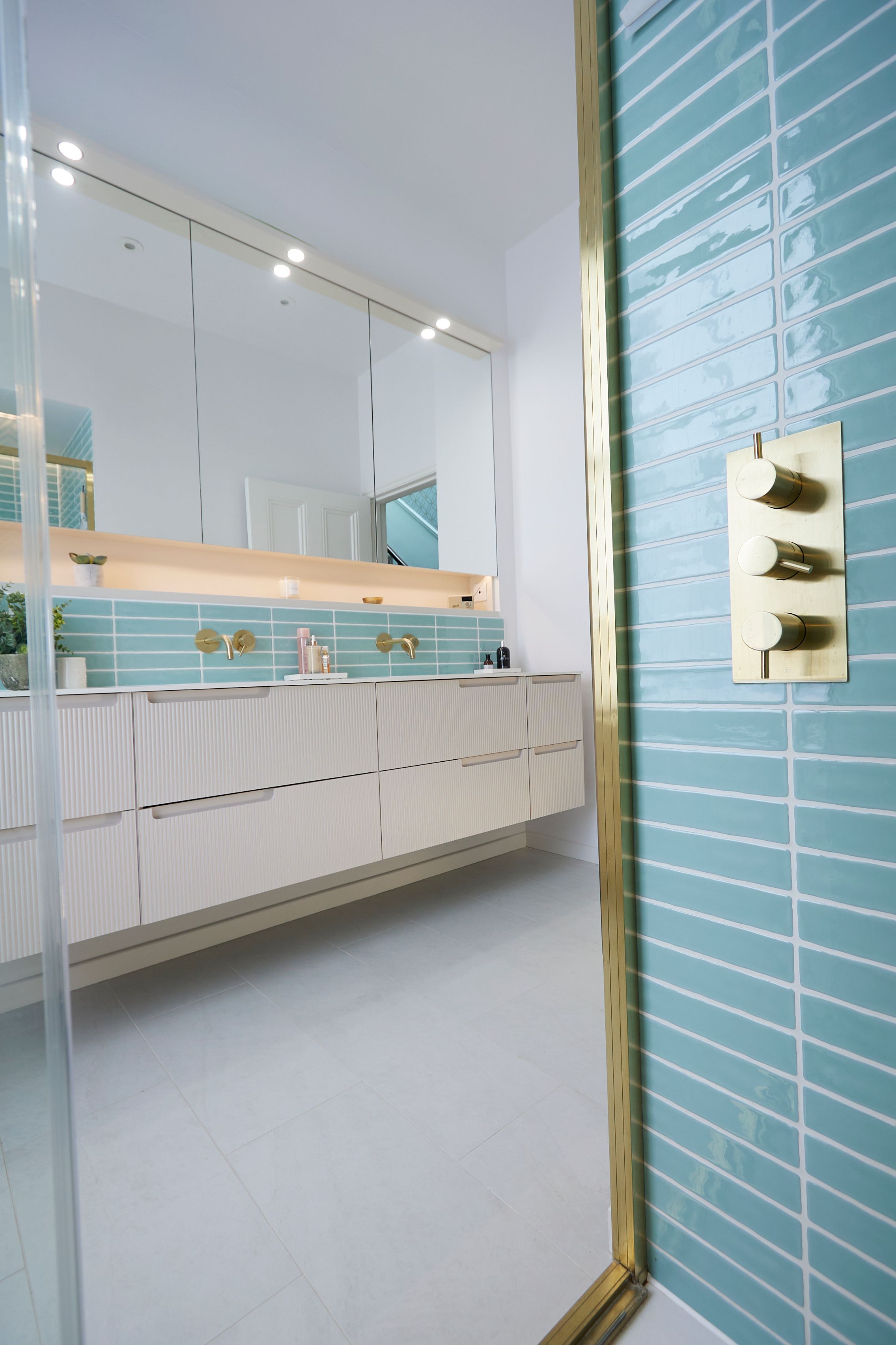 Modern bathroom interior featuring a sleek white vanity with ribbed texture, elegant gold fixtures, and a vibrant turquoise tiled wall. Mirrors above the sink enhance the space, while soft lighting creates a calming atmosphere.
