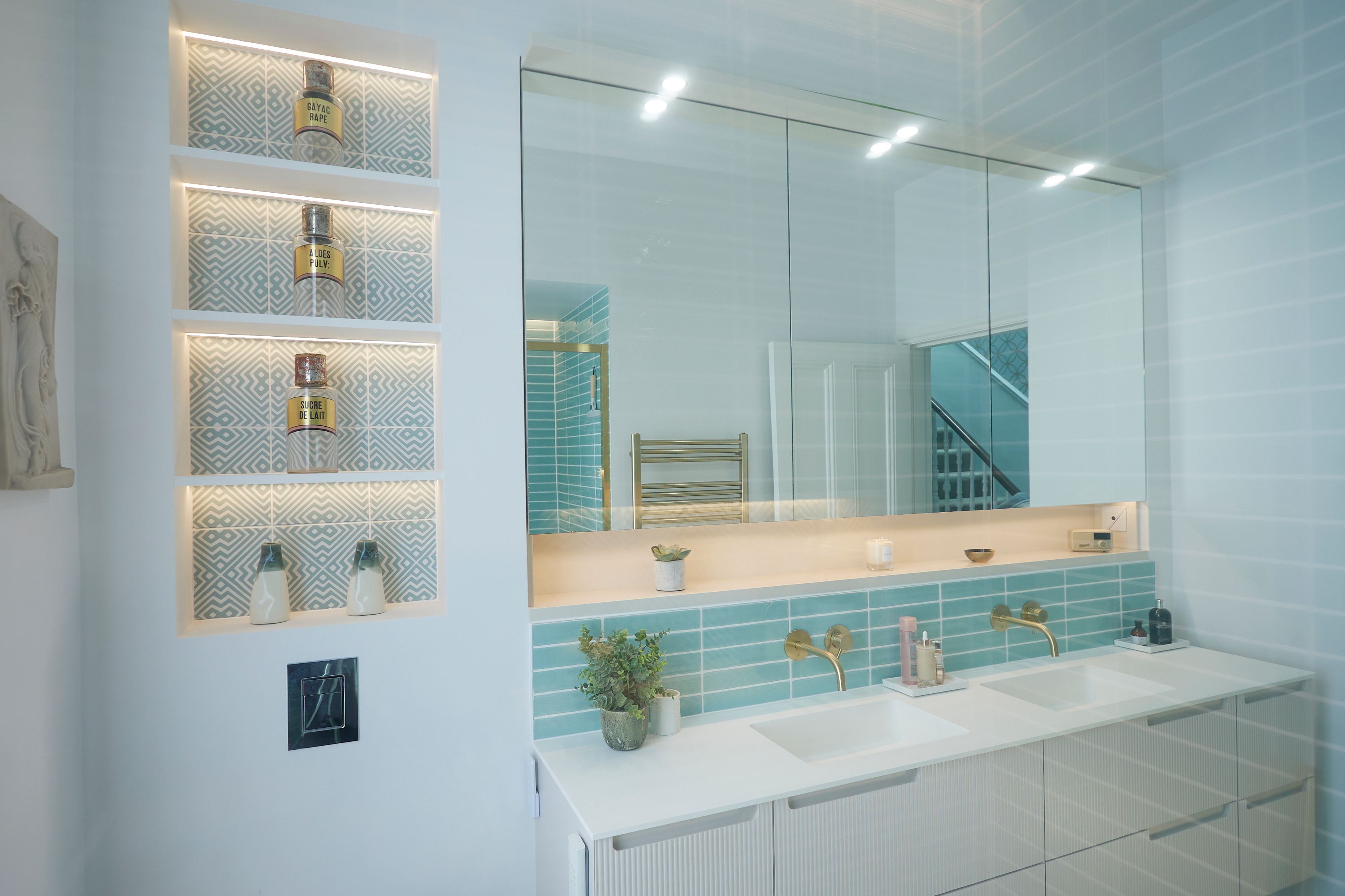 Modern bathroom interior featuring a double vanity with stylish gold taps, teal tiled backsplash, illuminated shelving displaying decorative bottles, and a large mirror. The design combines contemporary aesthetics with functional storage solutions.