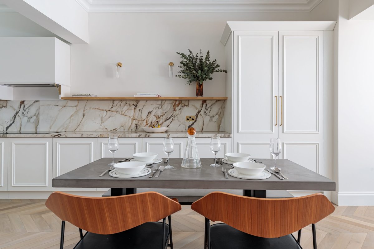 Modern dining area featuring a concrete table set for a meal with white bowls, cutlery, and glasses. The backdrop includes a stylish marble splashback and a wooden shelf adorned with a vase of greenery. An Arch KBB team member is present, contributing to the inviting atmosphere of the space.