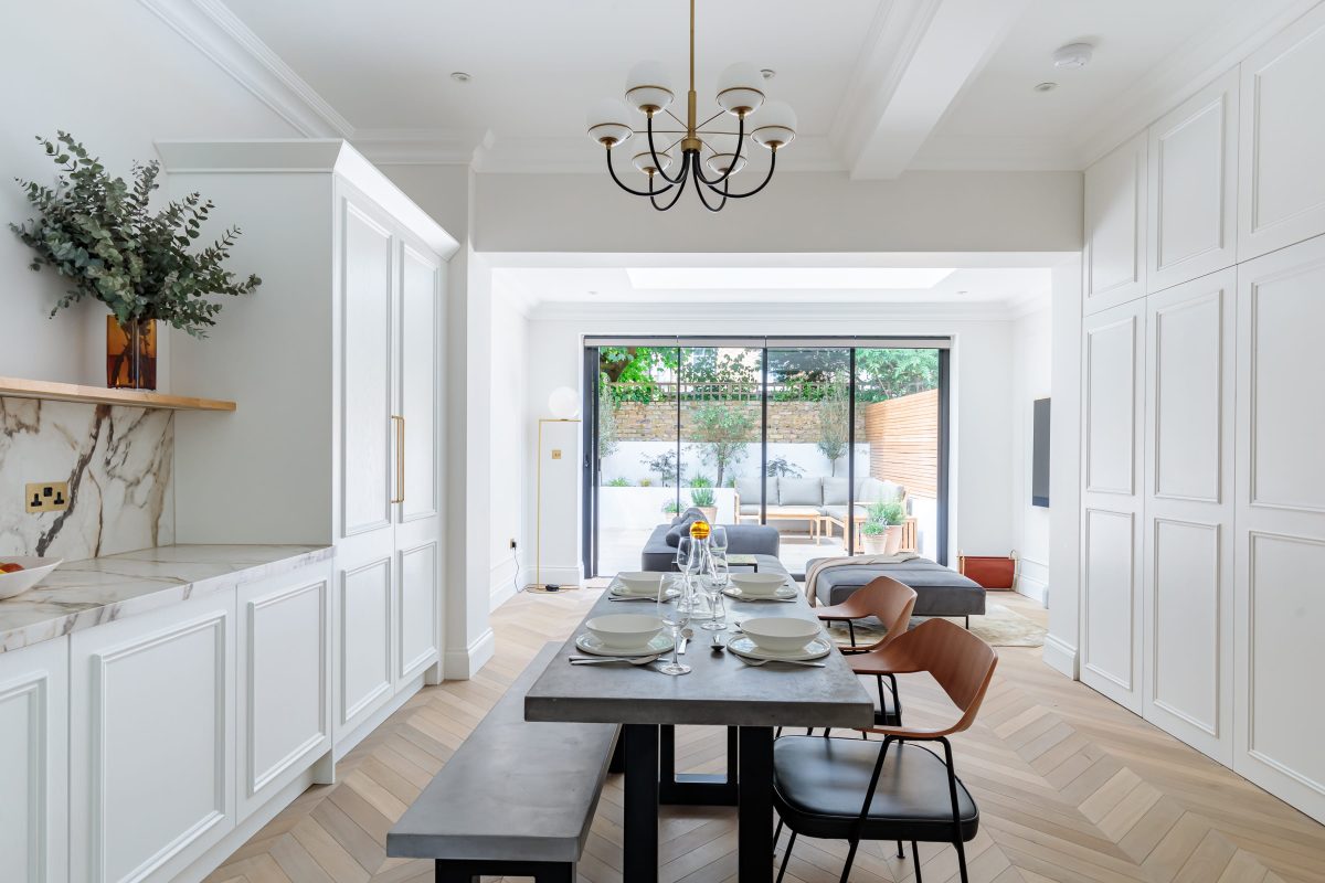 Modern dining area featuring a concrete table set with plates and glassware, surrounded by stylish chairs. The space includes elegant cabinetry with a marble countertop, and large glass doors leading to a bright outdoor area with seating and greenery. An Arch KBB team member is present, contributing to the welcoming atmosphere of the room.