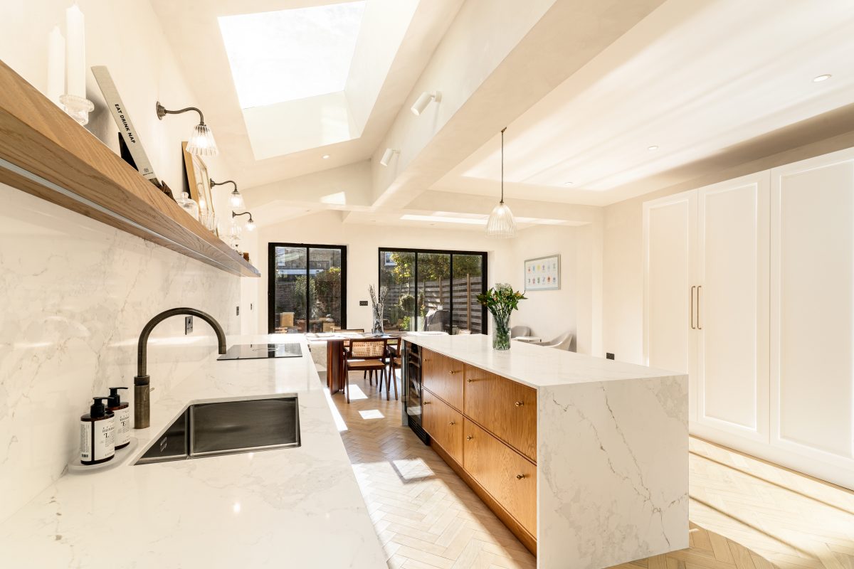 Modern kitchen interior featuring a marble countertop, wooden cabinetry, and large windows that allow natural light to illuminate the space. An Arch KBB team member is present, showcasing the stylish design elements, including pendant lighting and a sleek sink. The room has a warm and inviting atmosphere, ideal for both cooking and entertaining.