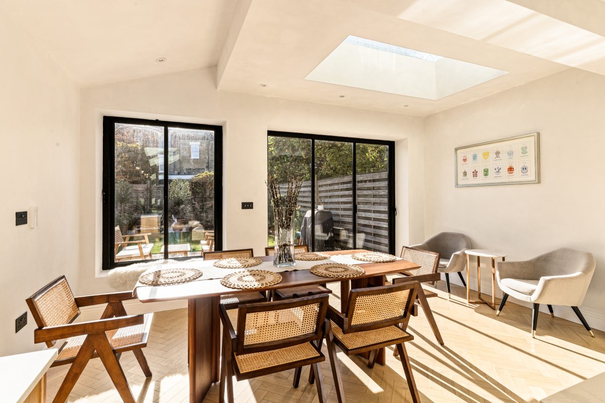 Bright and airy dining area featuring a wooden table set with woven placemats, surrounded by stylish chairs. Large windows provide views of a garden, while a skylight enhances natural light. An Arch KBB team member is present, contributing to the inviting atmosphere of the space.