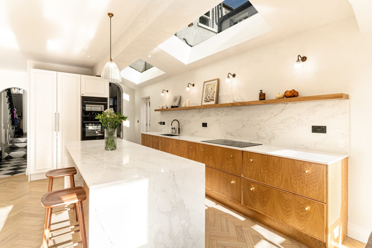 Modern kitchen interior featuring a spacious marble island, wooden cabinetry, and elegant lighting. The design includes skylights that enhance natural light, complemented by a vase of fresh flowers on the island. An Arch KBB team member is present, showcasing the kitchen's inviting atmosphere and contemporary style.