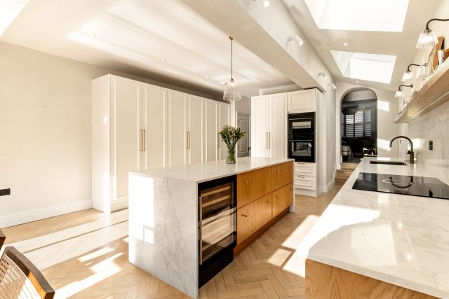 Bright and modern kitchen featuring a large central island with a marble top and wooden cabinetry. The space includes sleek white storage units, built-in appliances, and a stylish sink. Natural light floods the room through skylights, highlighting the herringbone-patterned wooden floor. An Arch KBB team member is present, enhancing the kitchen's inviting atmosphere.