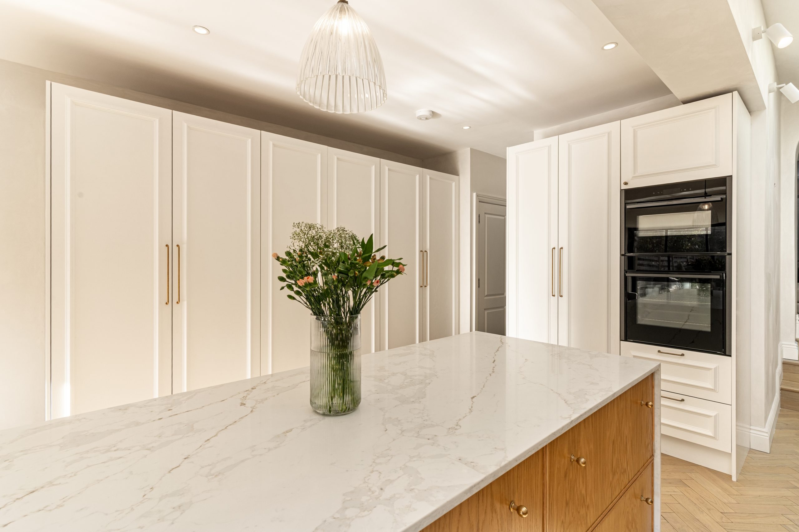 A modern kitchen featuring a marble island with a vase of fresh flowers, sleek white cabinetry, and an integrated oven. An Arch KBB team member is present, showcasing the elegant design and functionality of the space.