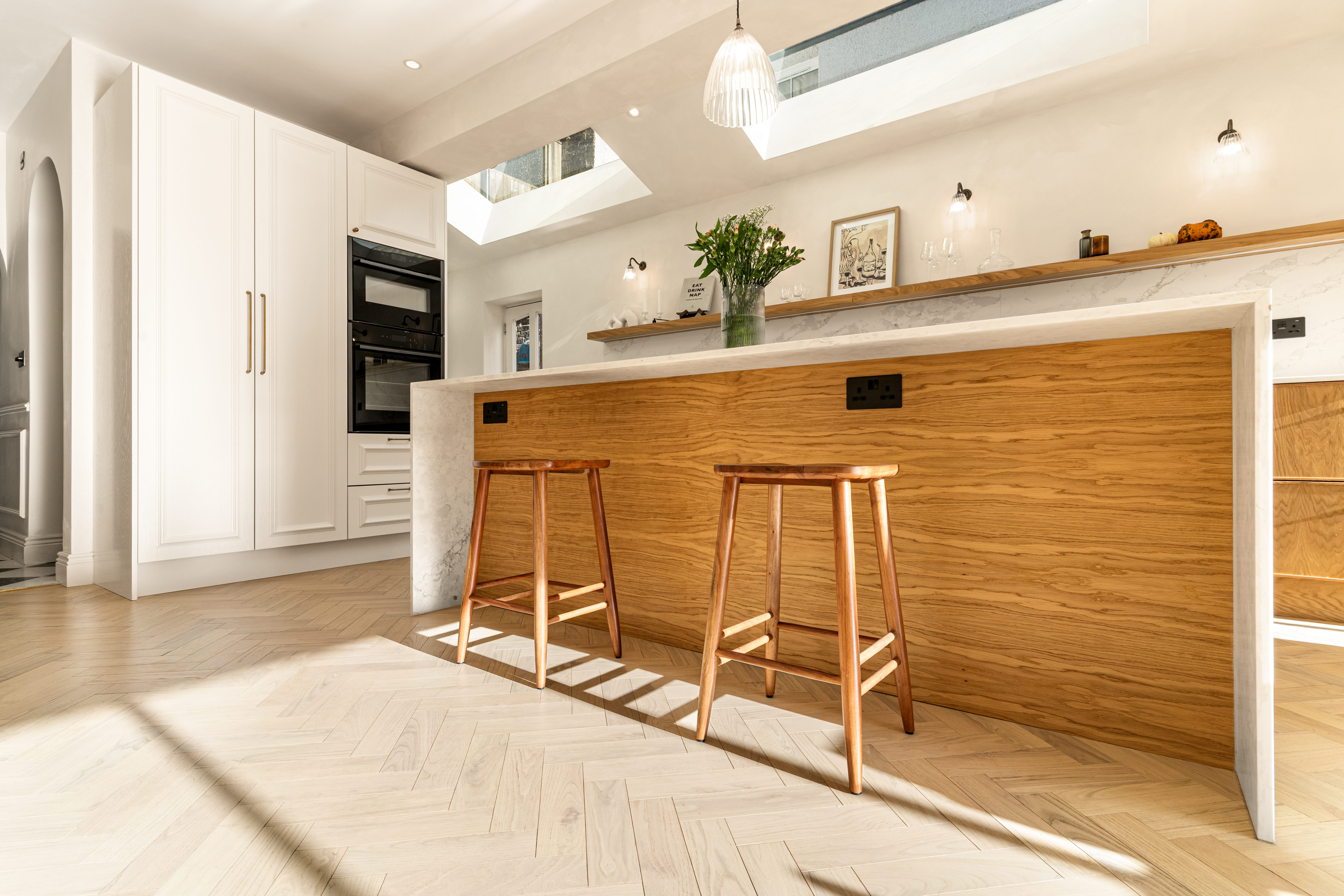 A modern kitchen featuring a wooden bar with two stools, sleek cabinetry, and a built-in oven. Natural light floods the space through skylights, highlighting the herringbone-patterned flooring and minimalist decor. An Arch KBB team member is present, adding to the inviting atmosphere.