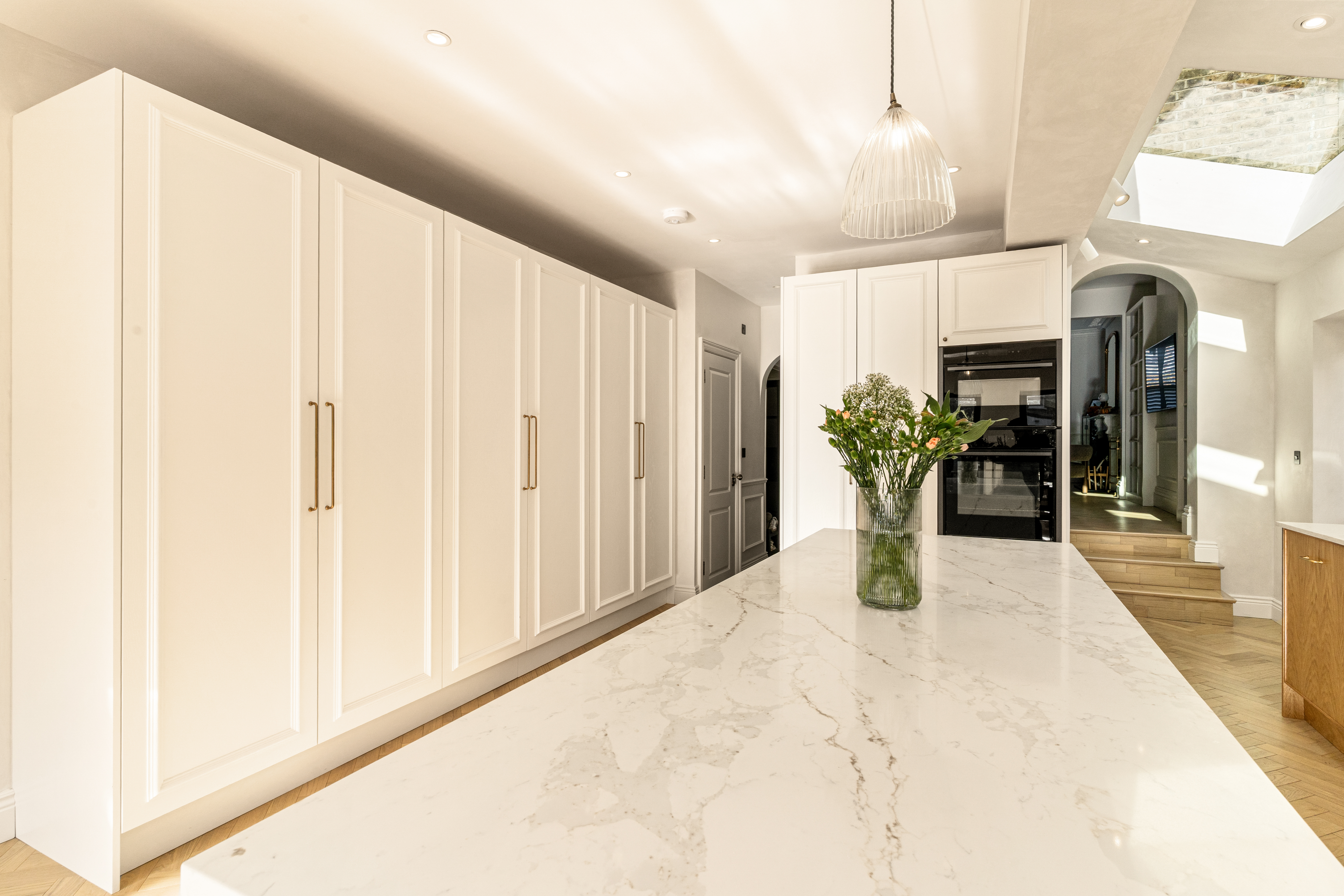 Modern kitchen design featuring a spacious marble countertop with a glass vase of flowers, complemented by sleek white cabinetry and warm wooden flooring. An Arch KBB team member is present, enhancing the aesthetic appeal of the contemporary interior. Natural light floods the space, highlighting the elegant finishes and creating an inviting atmosphere.