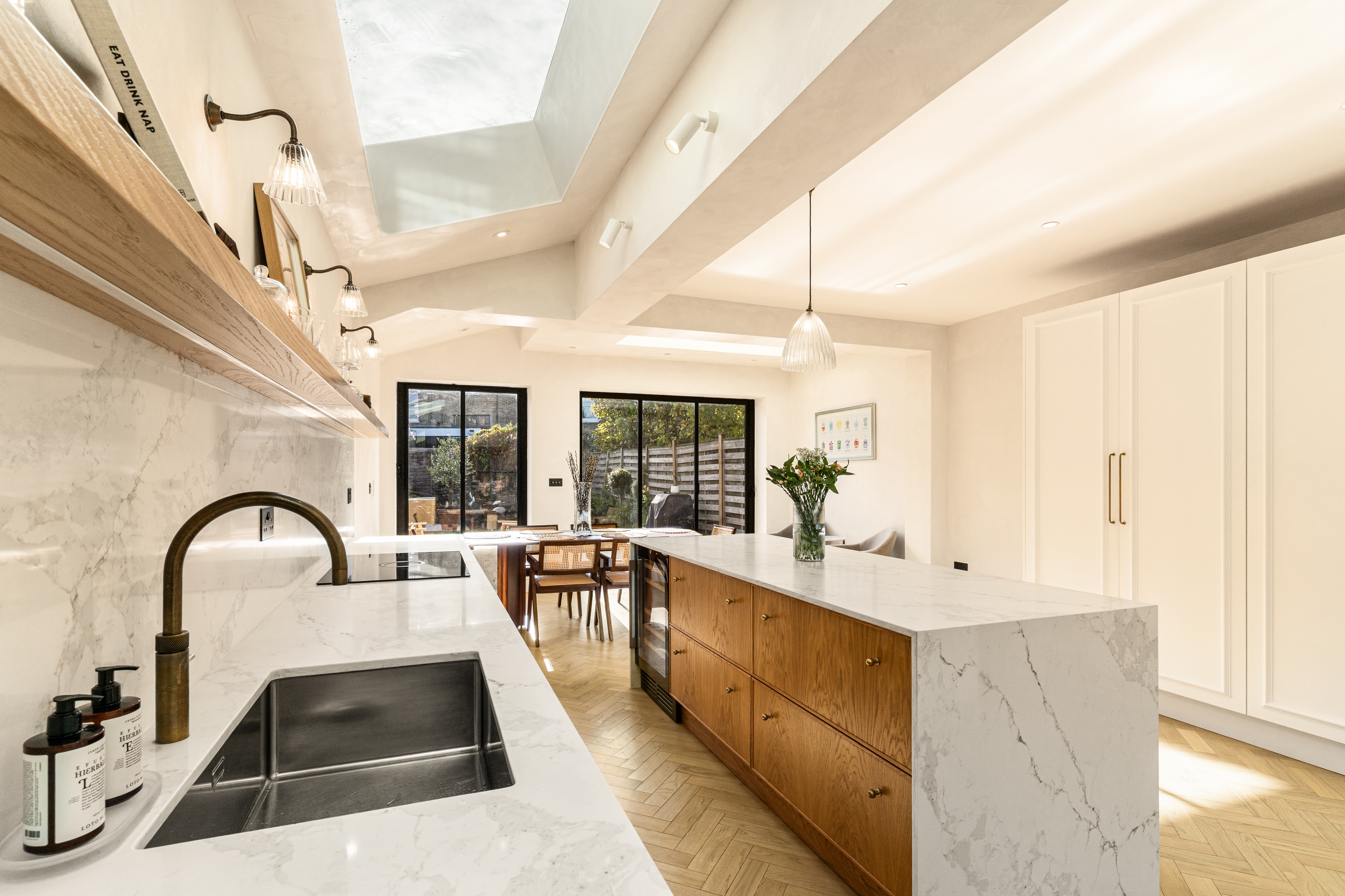 Modern kitchen interior featuring a stylish design with marble countertops, wooden cabinetry, and a skylight that enhances natural light. An Arch KBB team member is present, contributing to the warm and inviting atmosphere. The space includes a dining area with wooden chairs and a vase of flowers on the kitchen island, creating a harmonious blend of functionality and aesthetics.