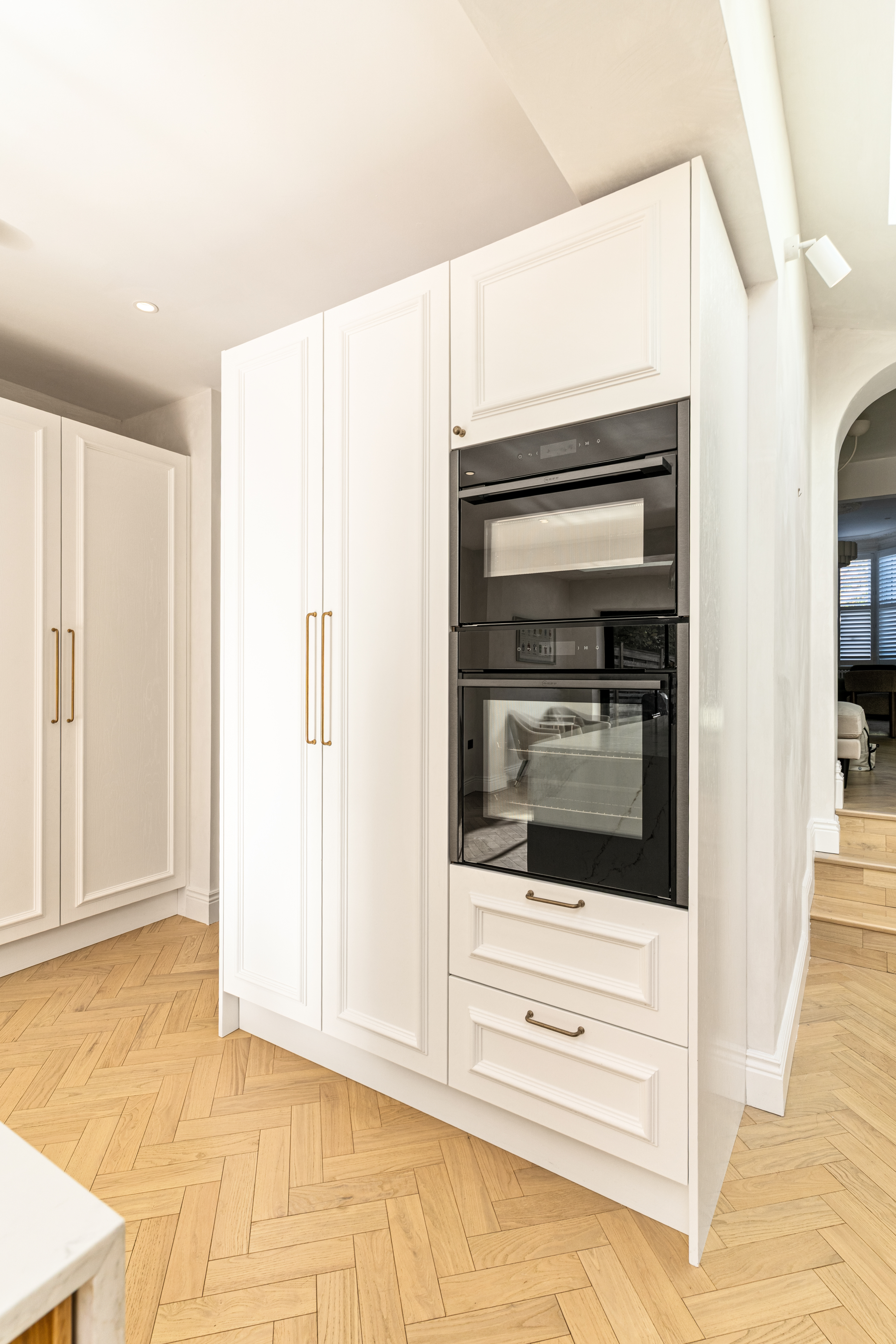 Modern kitchen featuring sleek white cabinetry and a built-in black oven. The herringbone patterned wooden floor adds warmth to the contemporary design. An Arch KBB team member is present, showcasing the stylish kitchen layout.