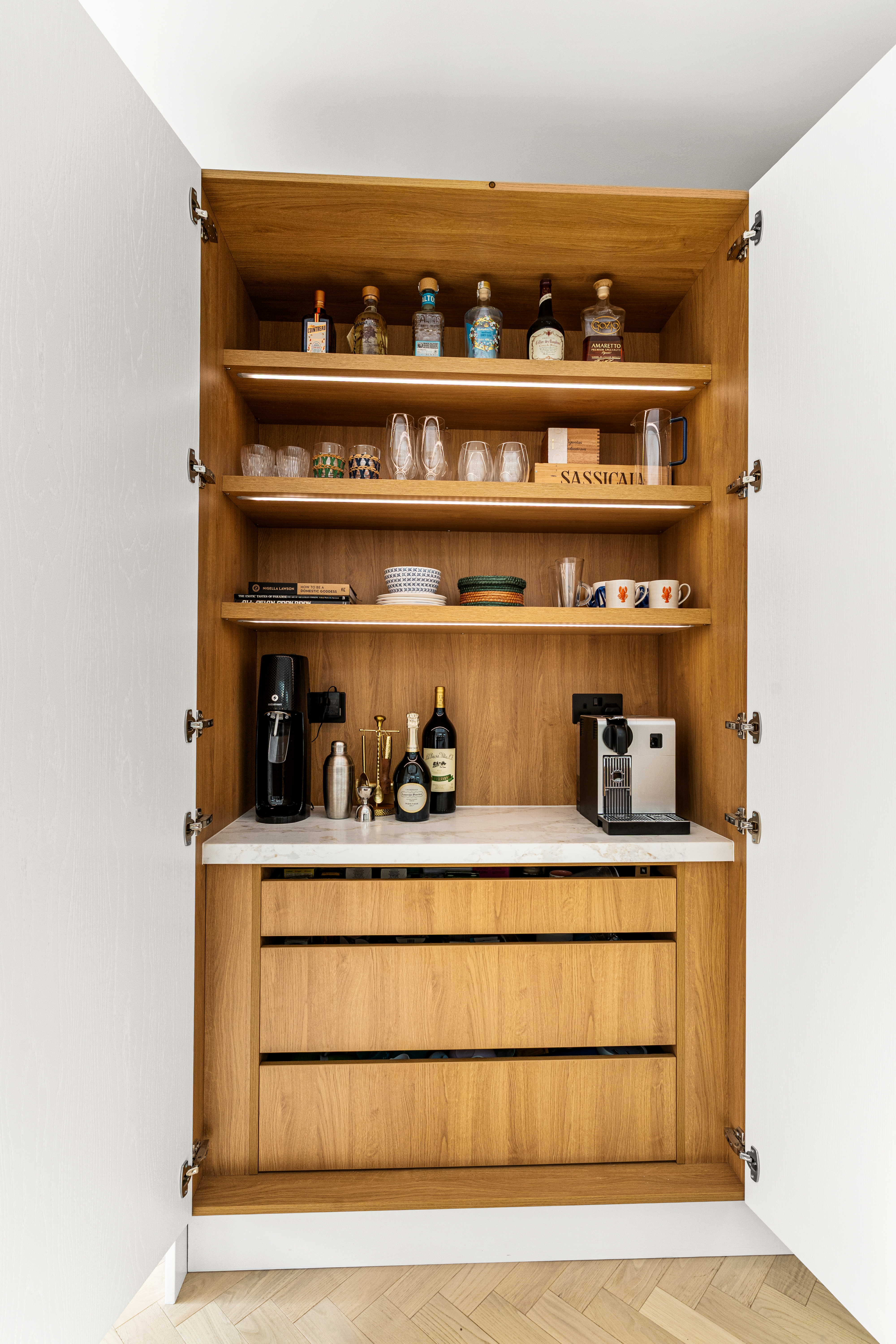 A stylish wooden bar cabinet with open shelves displaying various bottles of spirits, glassware, and decorative items. The cabinet features a sleek countertop with a coffee machine and wine bottles, complemented by three spacious drawers below. An Arch KBB team member is present, showcasing the elegant design and functionality of the space.