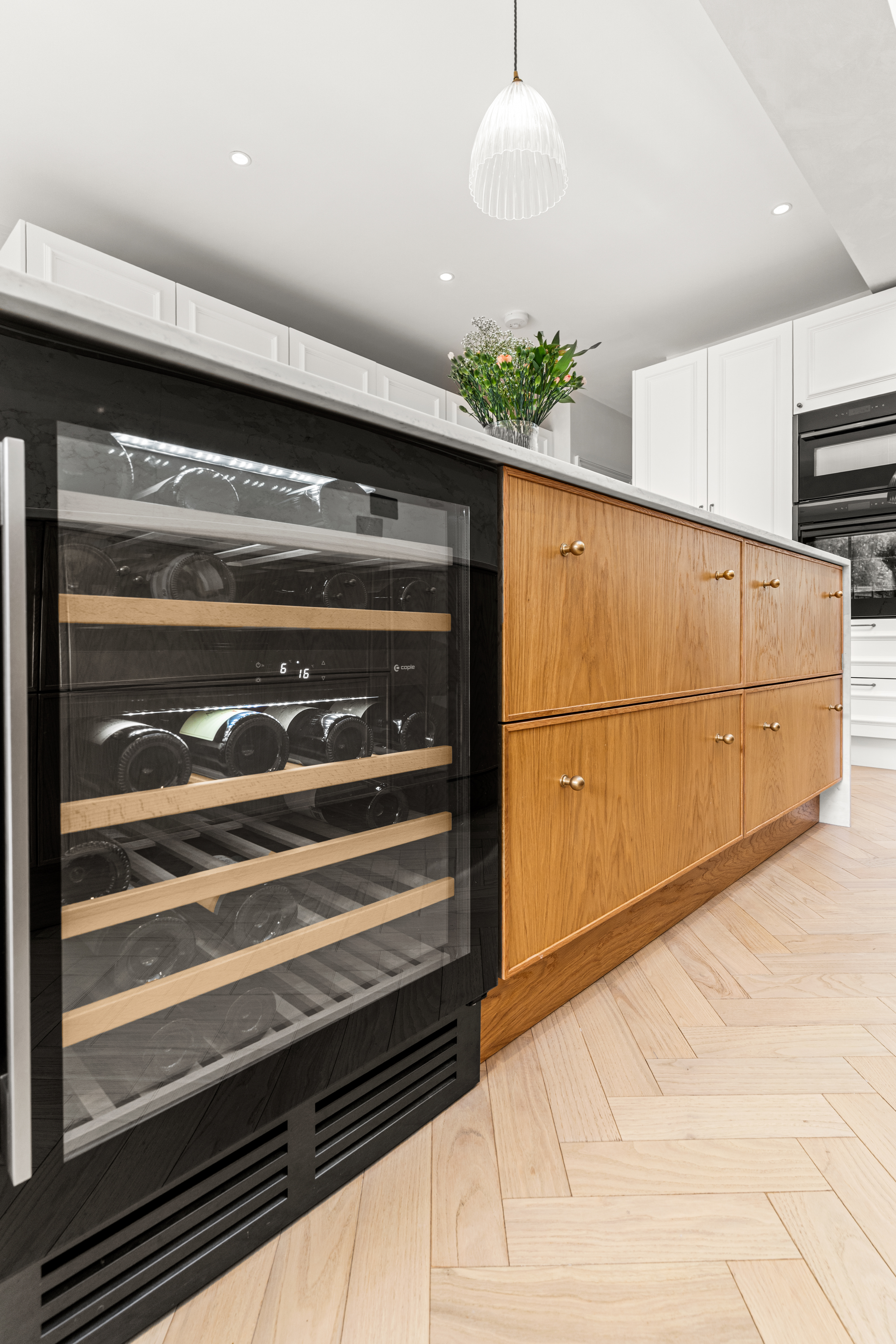 Modern kitchen featuring a stylish wine cooler integrated into the cabinetry, with wooden drawers and a sleek countertop. An Arch KBB team member is present, enhancing the design space. Natural light highlights the elegant finishes and herringbone flooring.