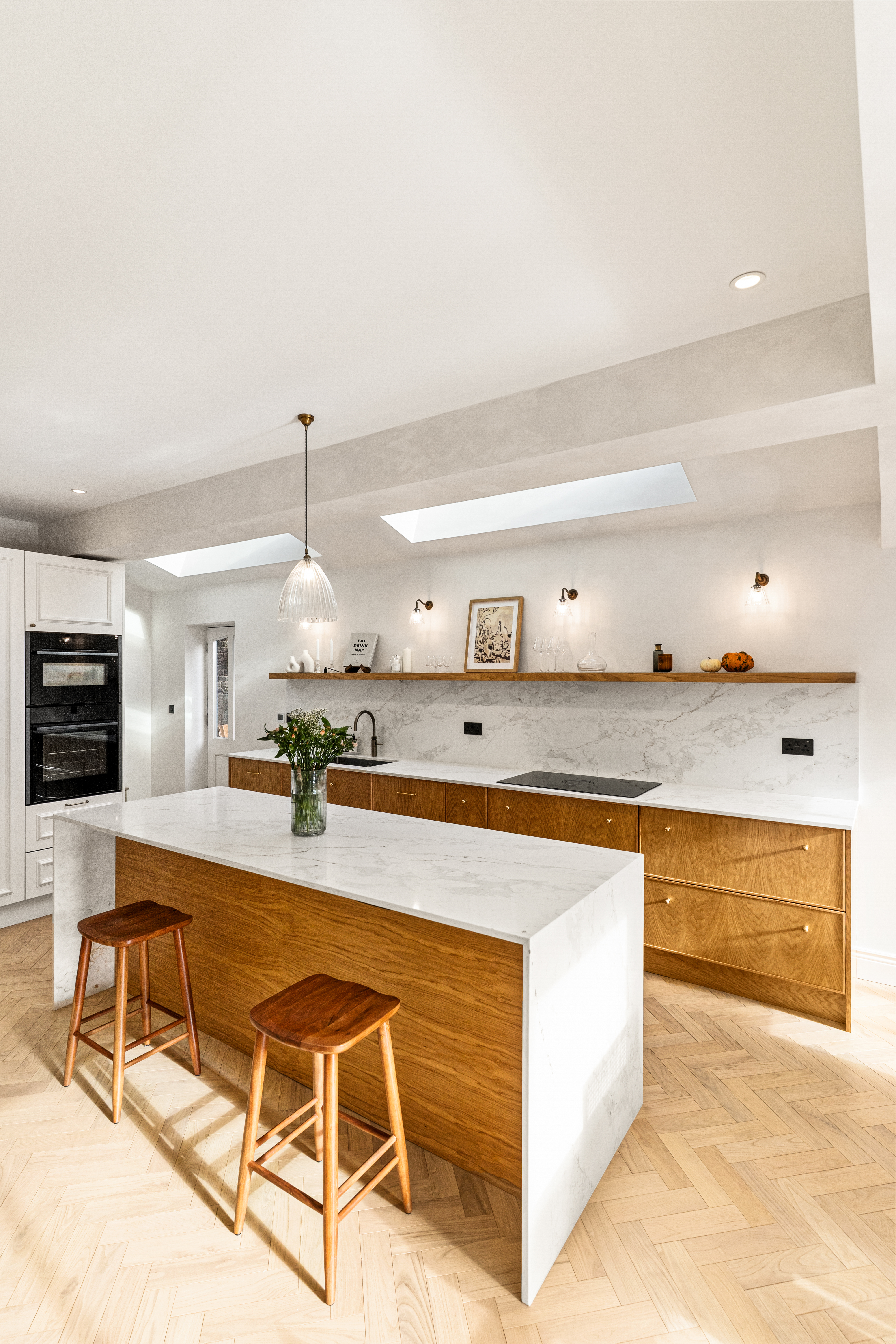 Modern kitchen design featuring a spacious island with a marble countertop and wooden cabinetry. The room is illuminated by natural light from skylights and stylish pendant lighting. An Arch KBB team member is present, adding to the inviting atmosphere. The decor includes minimalist shelving with decorative items, enhancing the contemporary aesthetic.
