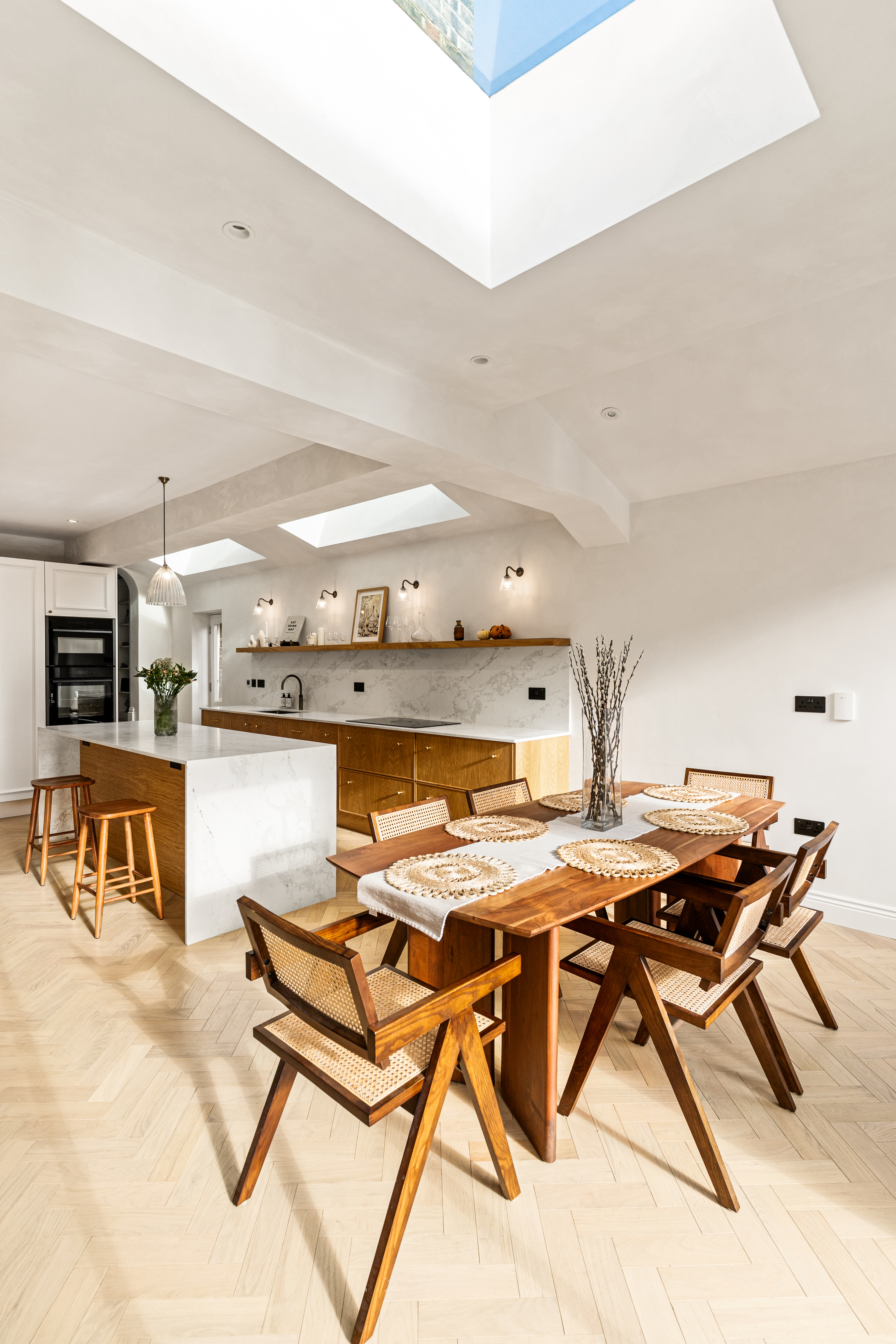 Modern kitchen and dining area featuring a minimalist design with wooden furniture, a marble-topped island, and natural light streaming through skylights. The dining table is set with woven placemats and decorative branches in a vase, creating an inviting atmosphere. An Arch KBB team member can be seen in the background, contributing to the space's aesthetic appeal.