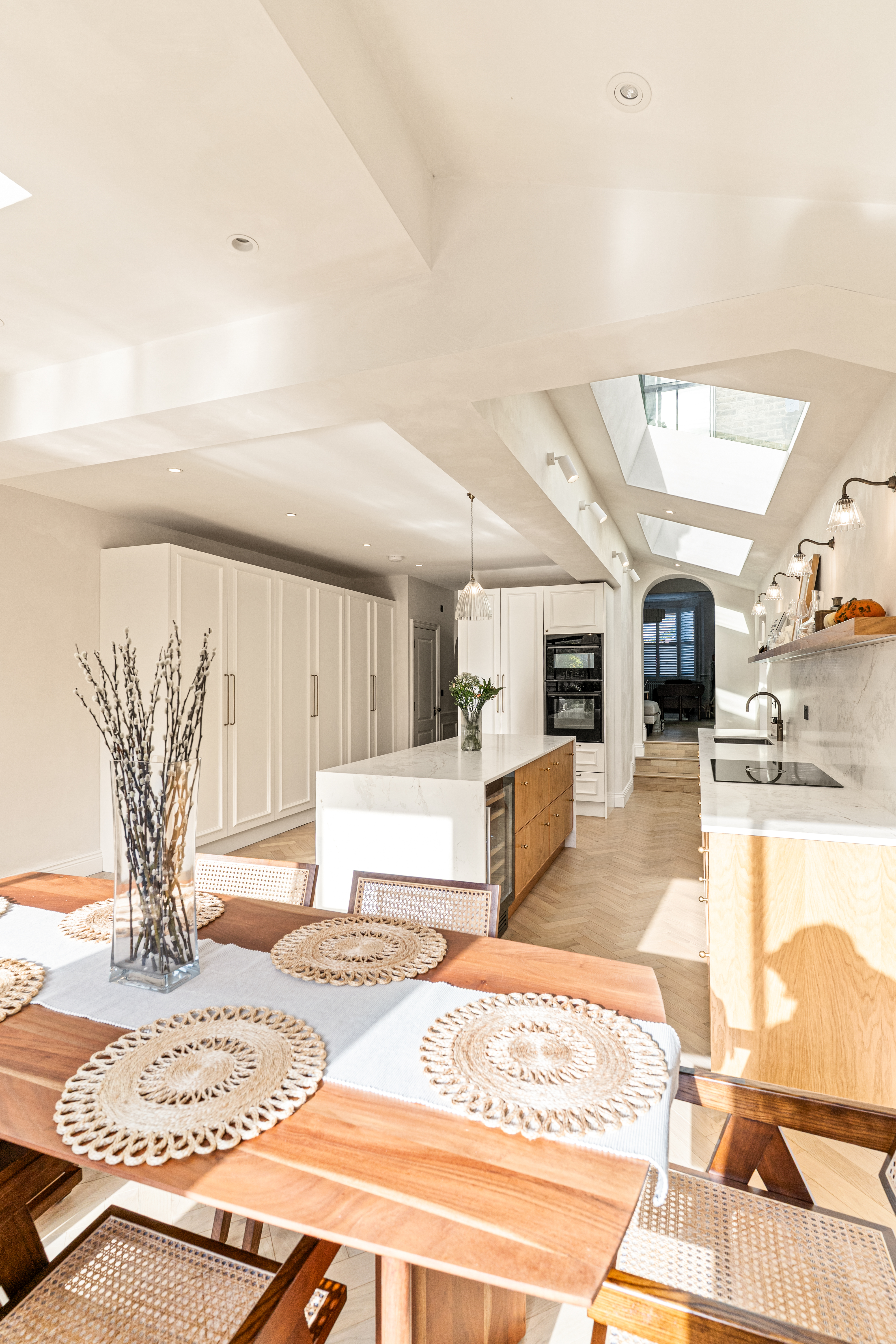 Bright and modern kitchen featuring a spacious island with a marble countertop, wooden cabinetry, and sleek appliances. A dining table adorned with decorative placemats sits in the foreground, complemented by a vase of branches. Natural light floods the space through skylights, enhancing the airy atmosphere. An Arch KBB team member is present, contributing to the design process.