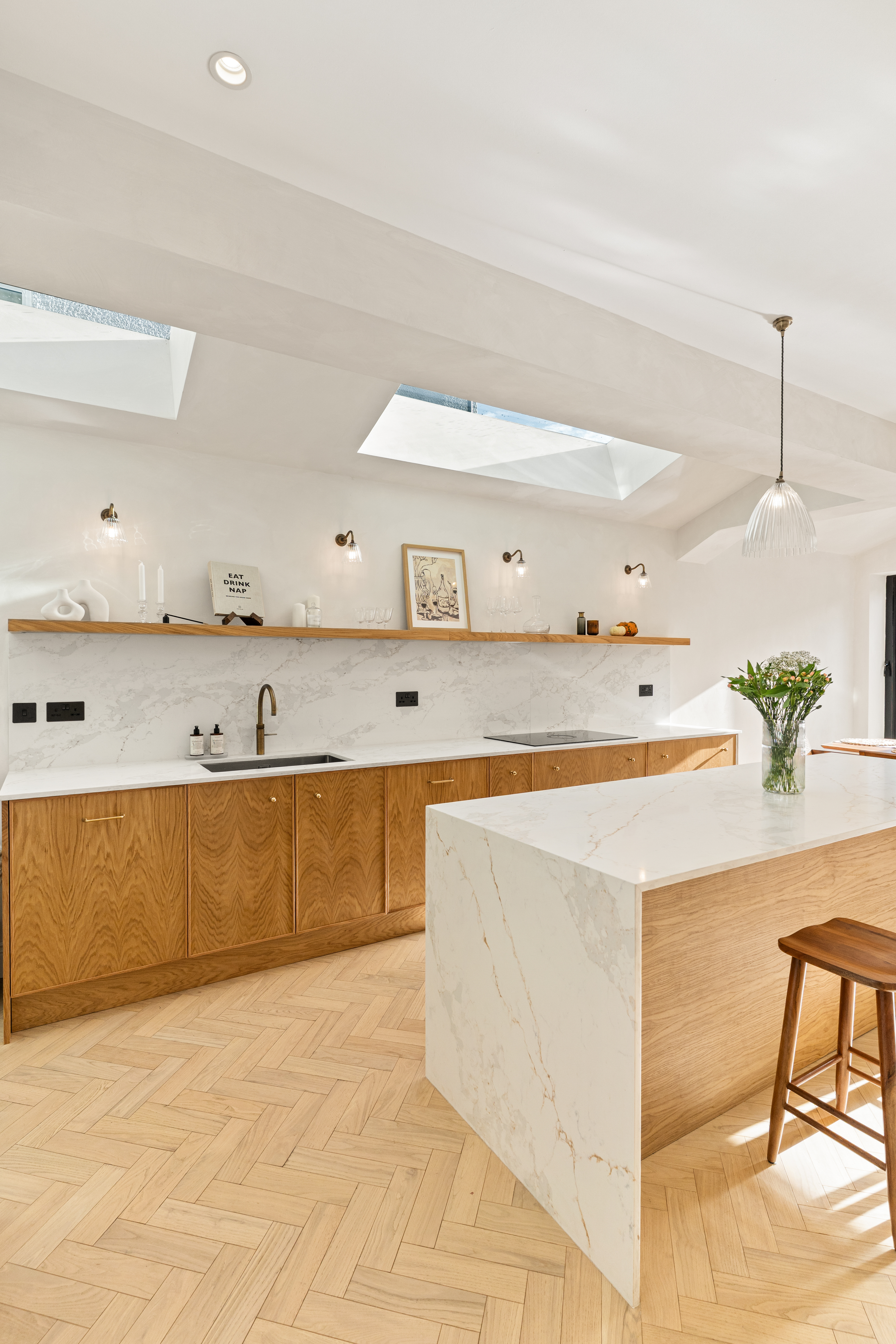 Modern kitchen design featuring a spacious layout with wooden cabinetry, a marble countertop, and stylish pendant lighting. The room is illuminated by natural light from skylights, showcasing a clean aesthetic with decorative elements and a vase of flowers on the countertop. An Arch KBB team member is present, contributing to the design atmosphere.