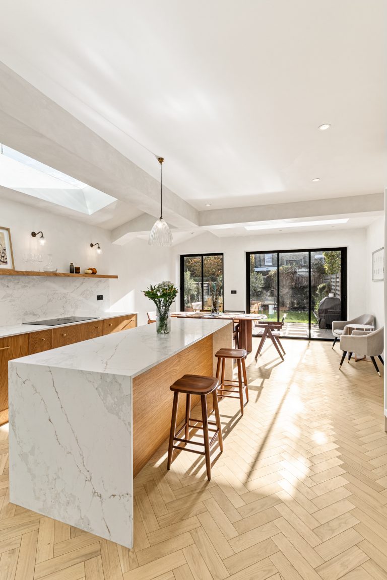 Bright and modern kitchen featuring a large island with a marble countertop, wooden cabinetry, and herringbone flooring. Natural light floods the space through skylights and large glass doors leading to a garden. An Arch KBB team member is present, enhancing the inviting atmosphere.