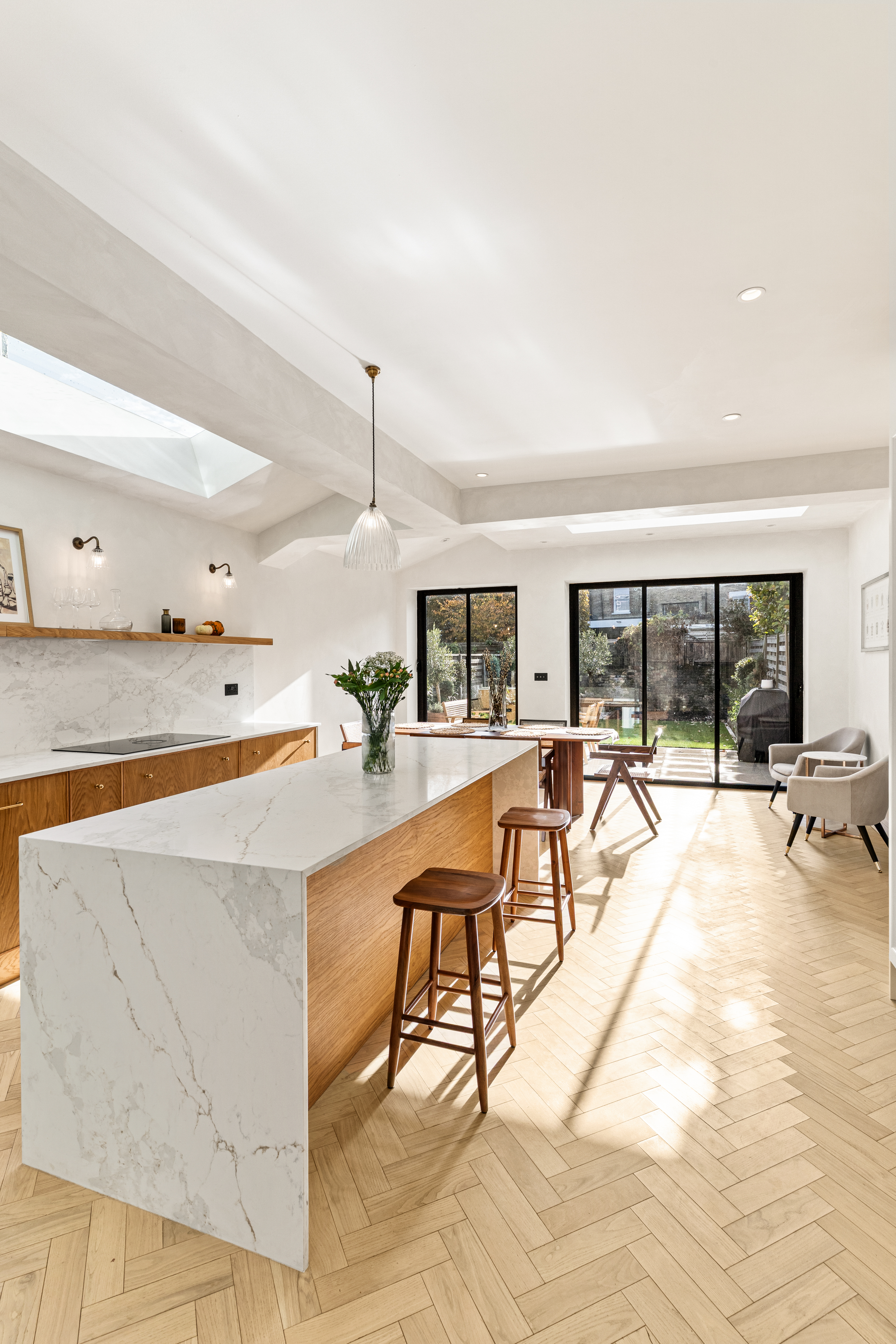 Bright and modern kitchen featuring a large island with a marble countertop, wooden cabinetry, and herringbone flooring. Natural light floods the space through skylights and large glass doors leading to a garden. An Arch KBB team member is present, enhancing the inviting atmosphere.